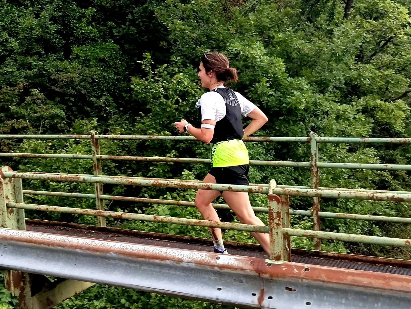 Une femme court sur un pont en métal avec des barrières rouillées devant une végétation dense.