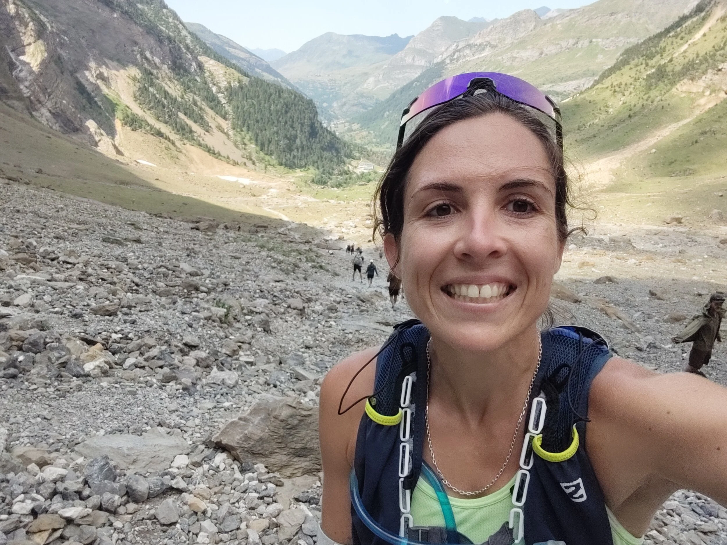 Femme souriante en trail dans un paysage de montagne rocheux avec des collines verdoyantes au fond