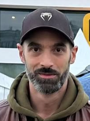 Un homme portant une casquette noire et un pull vert foncé, avec la barbe grise, devant un autobus.