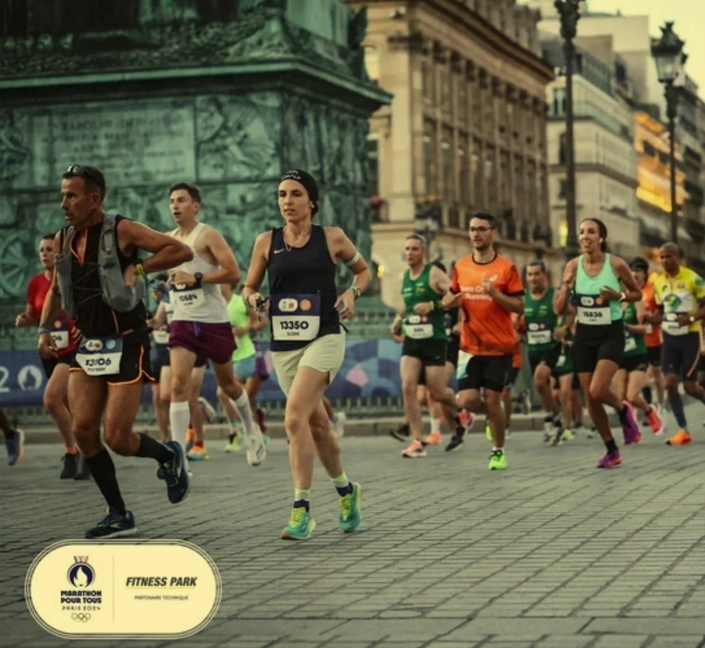 Des marathoniennes et marathoniens courent dans une rue urbaine avec des bâtiments historiques en arrière-plan, lors du Marathon pour Tous pour les JO 2024 de Paris.