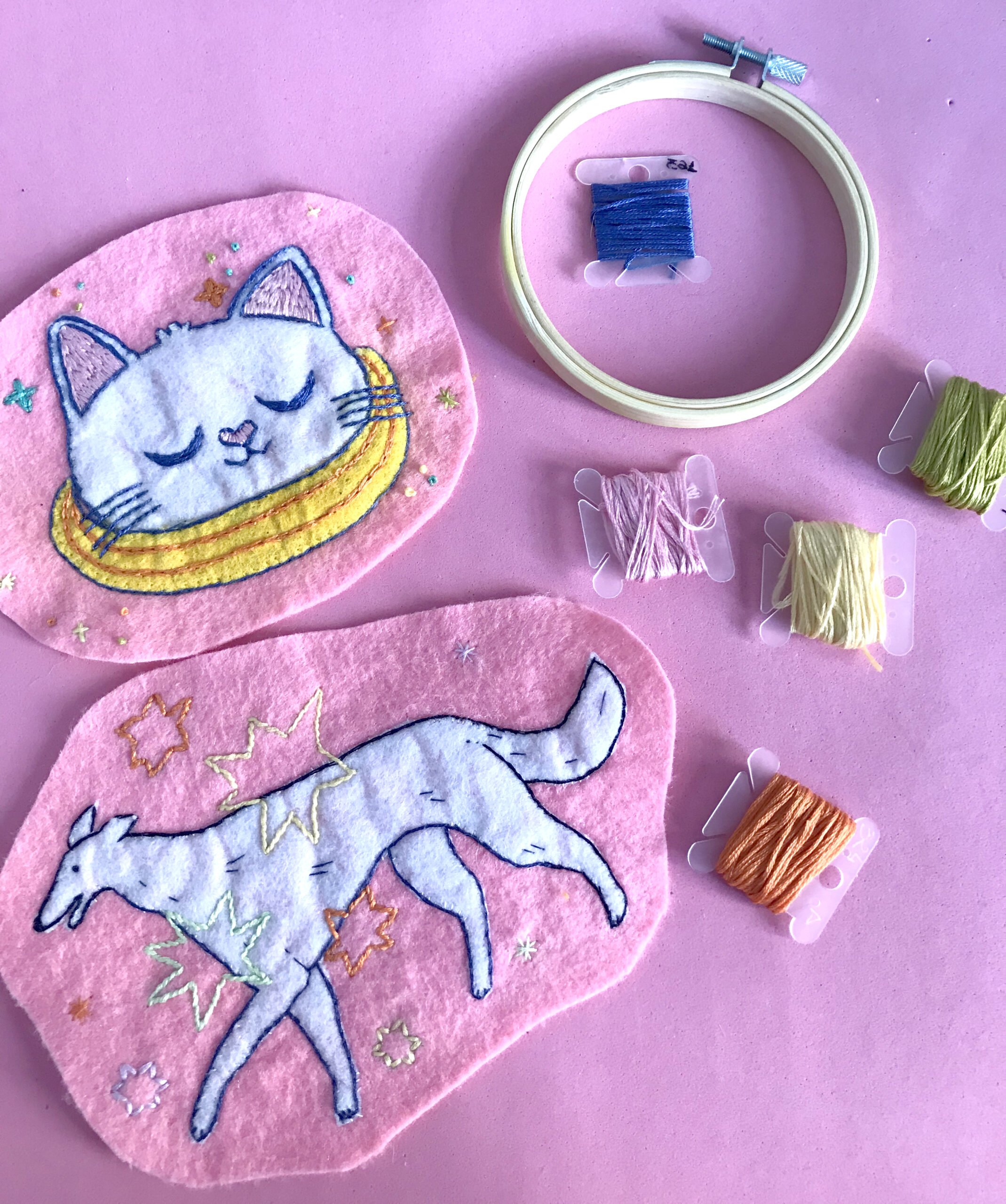 Dos piezas de tela con bordados de gato dormido y unicornio en fondo rosa, junto a carretes de hilo y un marco de bordado con hilo azul.