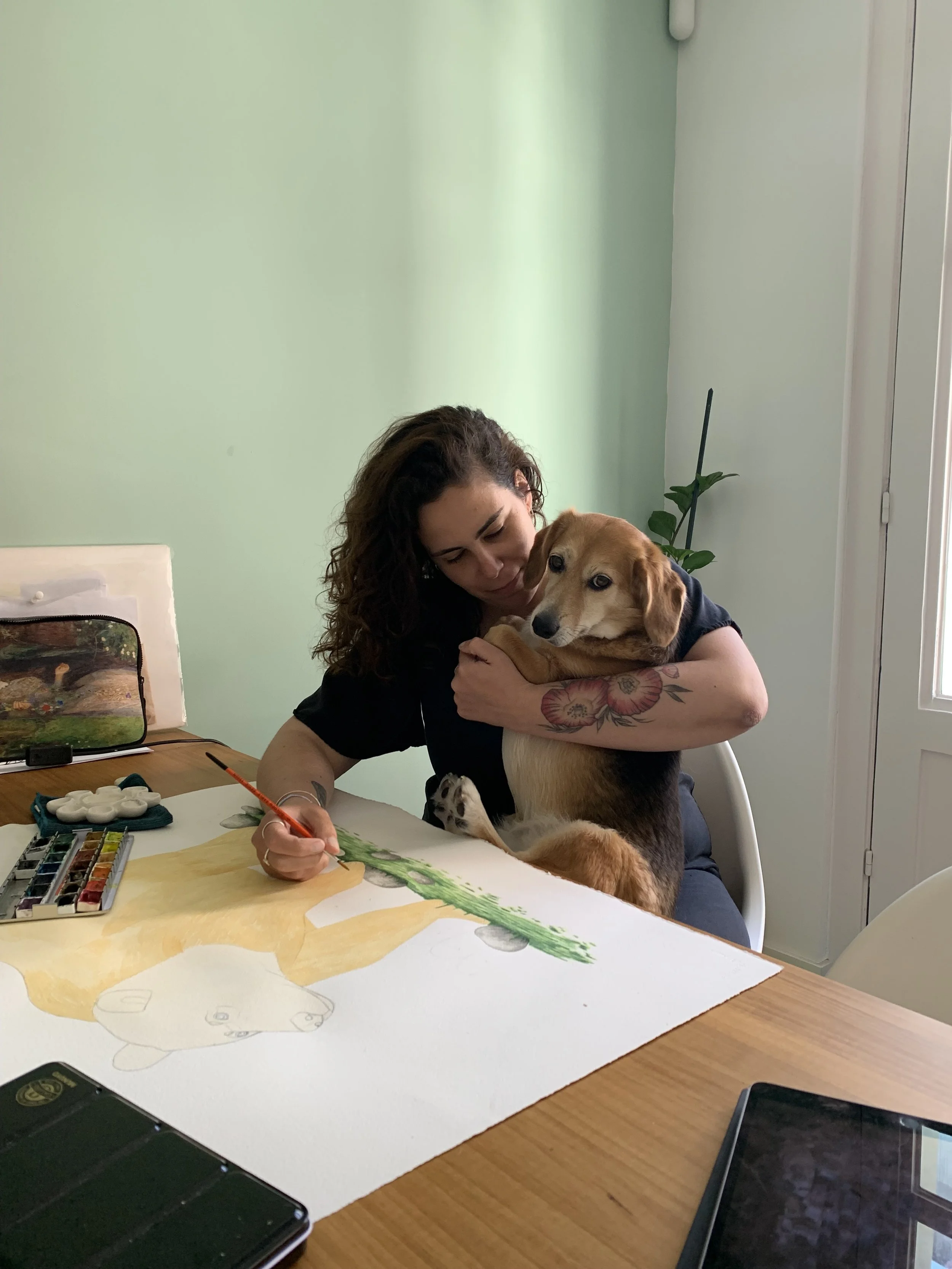 Una mujer con tatuajes en el brazo, pintando un cuadro en la mesa mientras un perro la abraza.