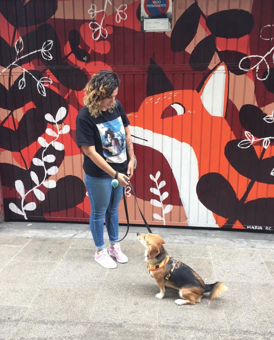 Una mujer con una camiseta negra y jeans azules está con un perro sentado en la acera, frente a un mural de un zorro en colores naranja, blanco, negro y marrón, decorado con hojas blancas en el fondo.