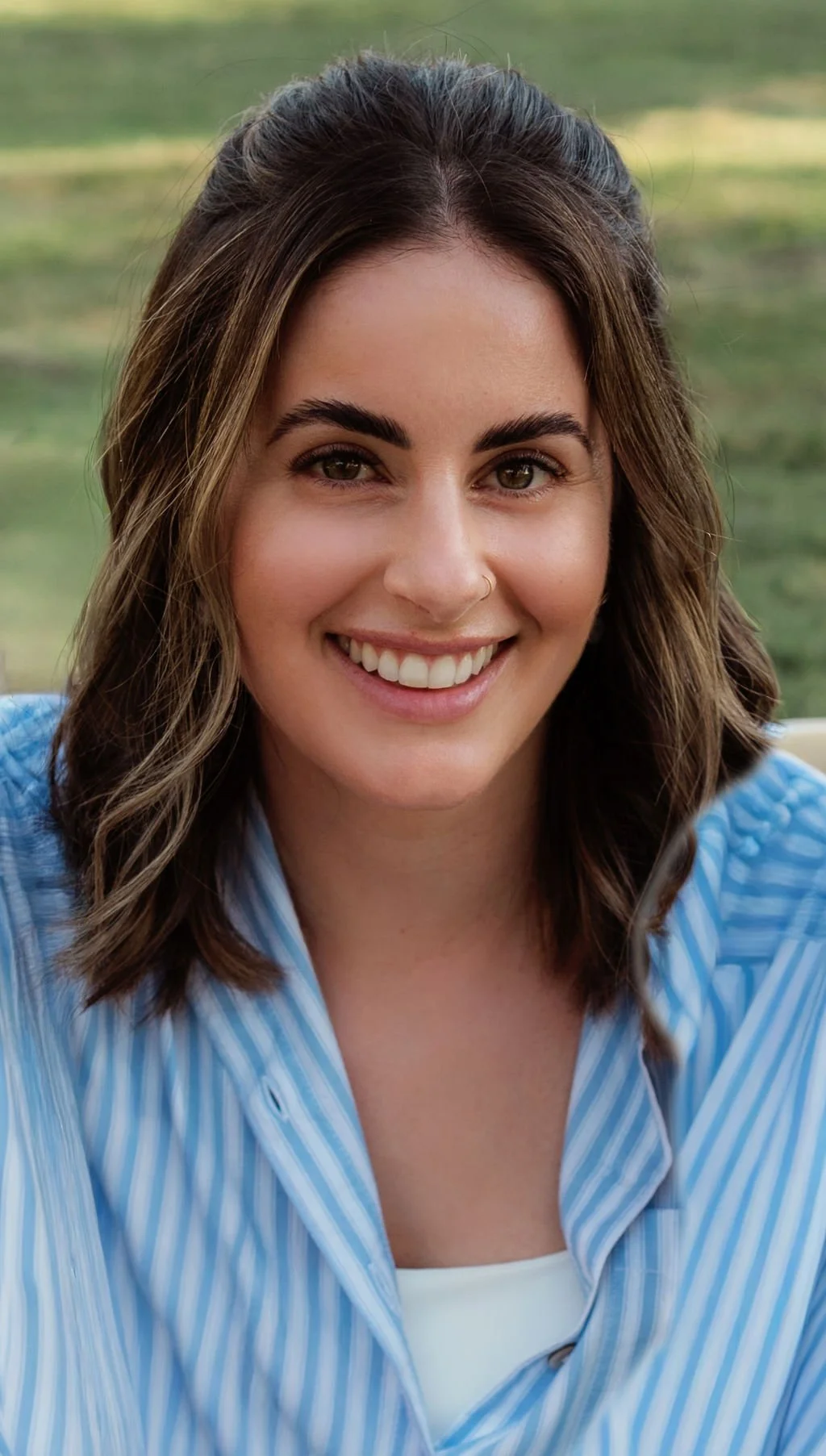 Close-up of smiling woman with shoulder-length brown hair, wearing a blue and white striped shirt outdoors.