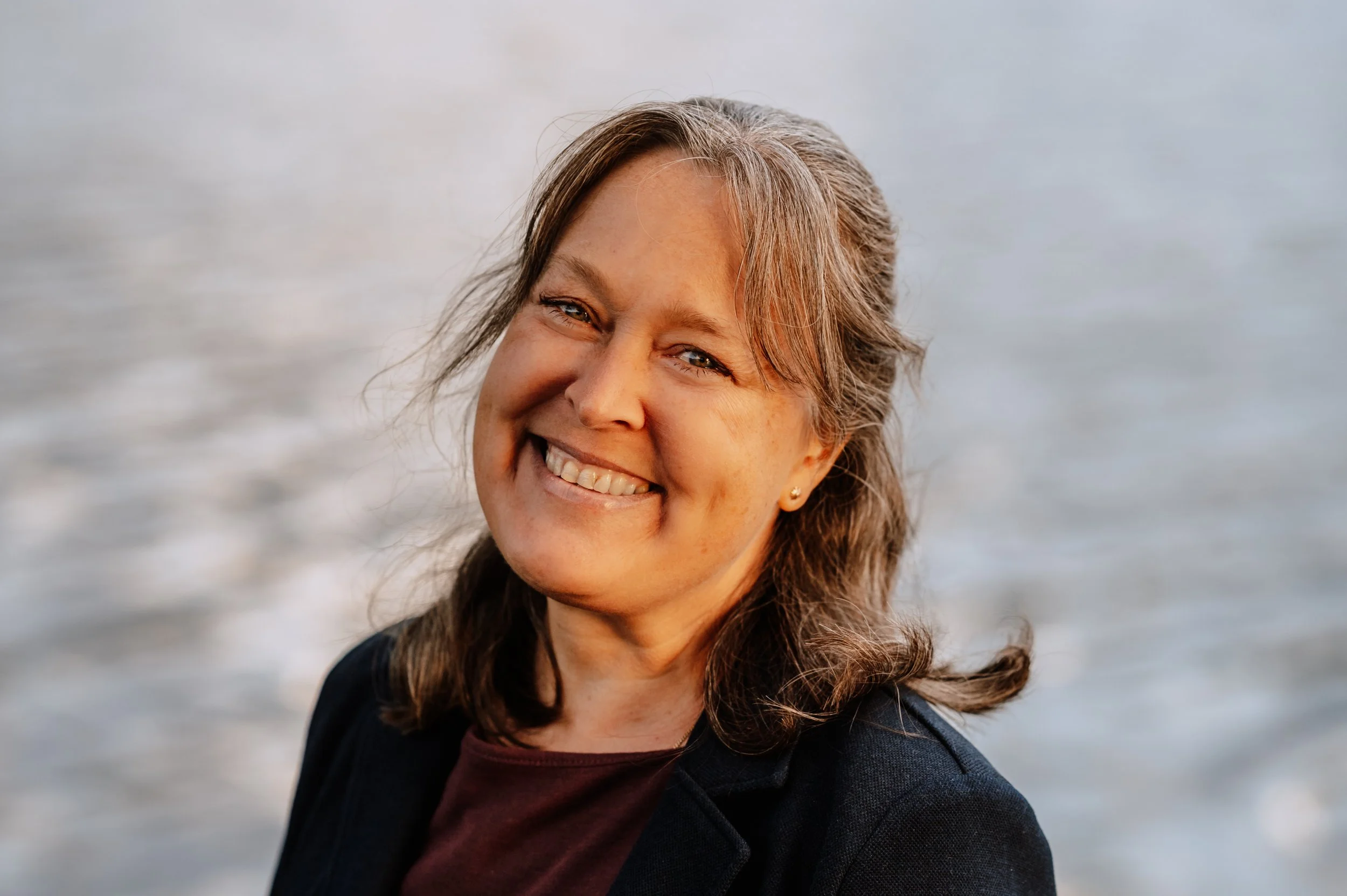 Lächelnde Frau mit braunen Haaren und schwarzen Jacke vor Wasser