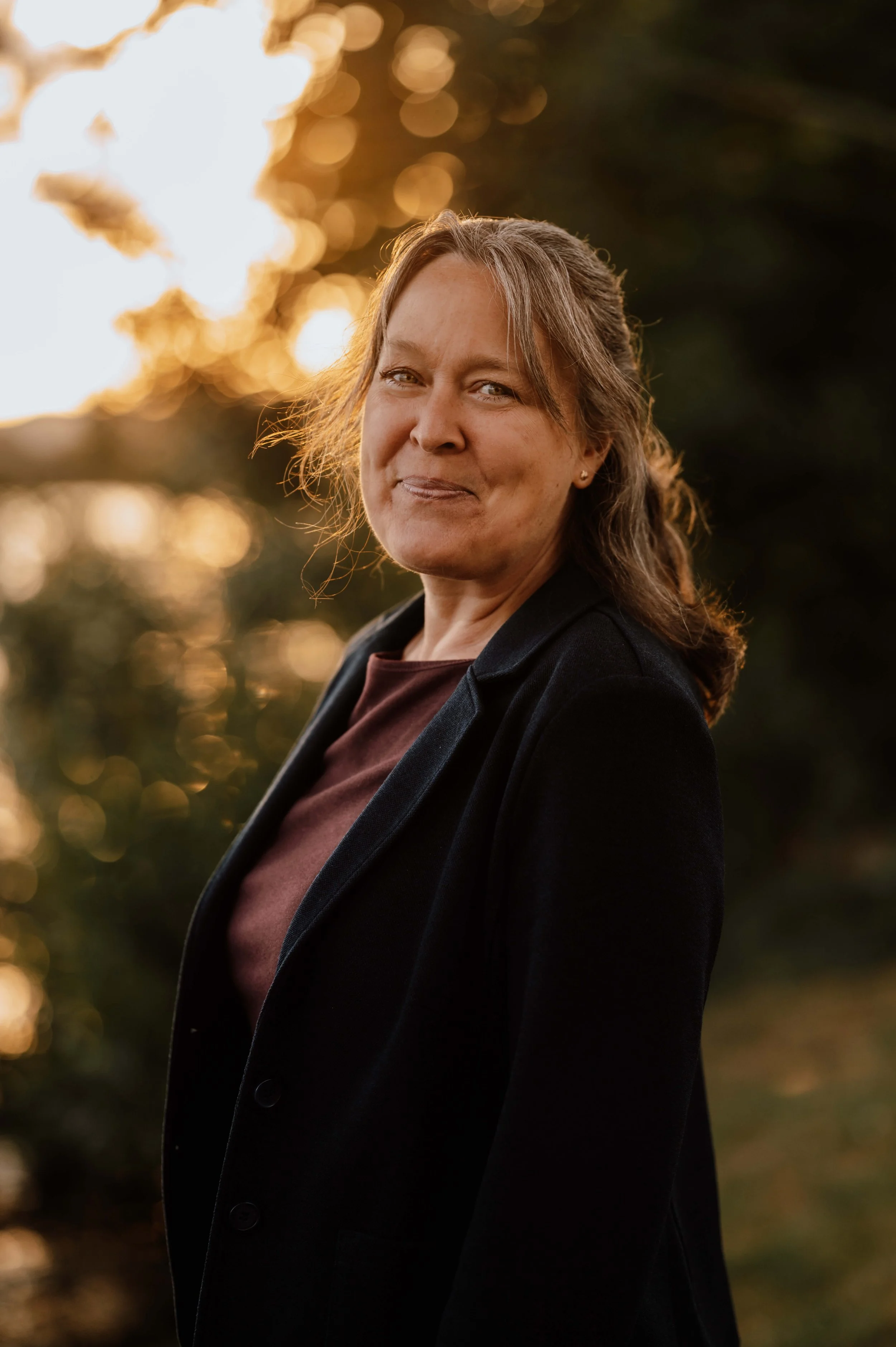 Porträt einer Frau mit braunen Haaren, die auf einem Hügel bei Sonnenuntergang steht, im schwarzen Blazer und mit einem warmen Lächeln, im Hintergrund verschwommene Bäume und goldene Lichteffekte.