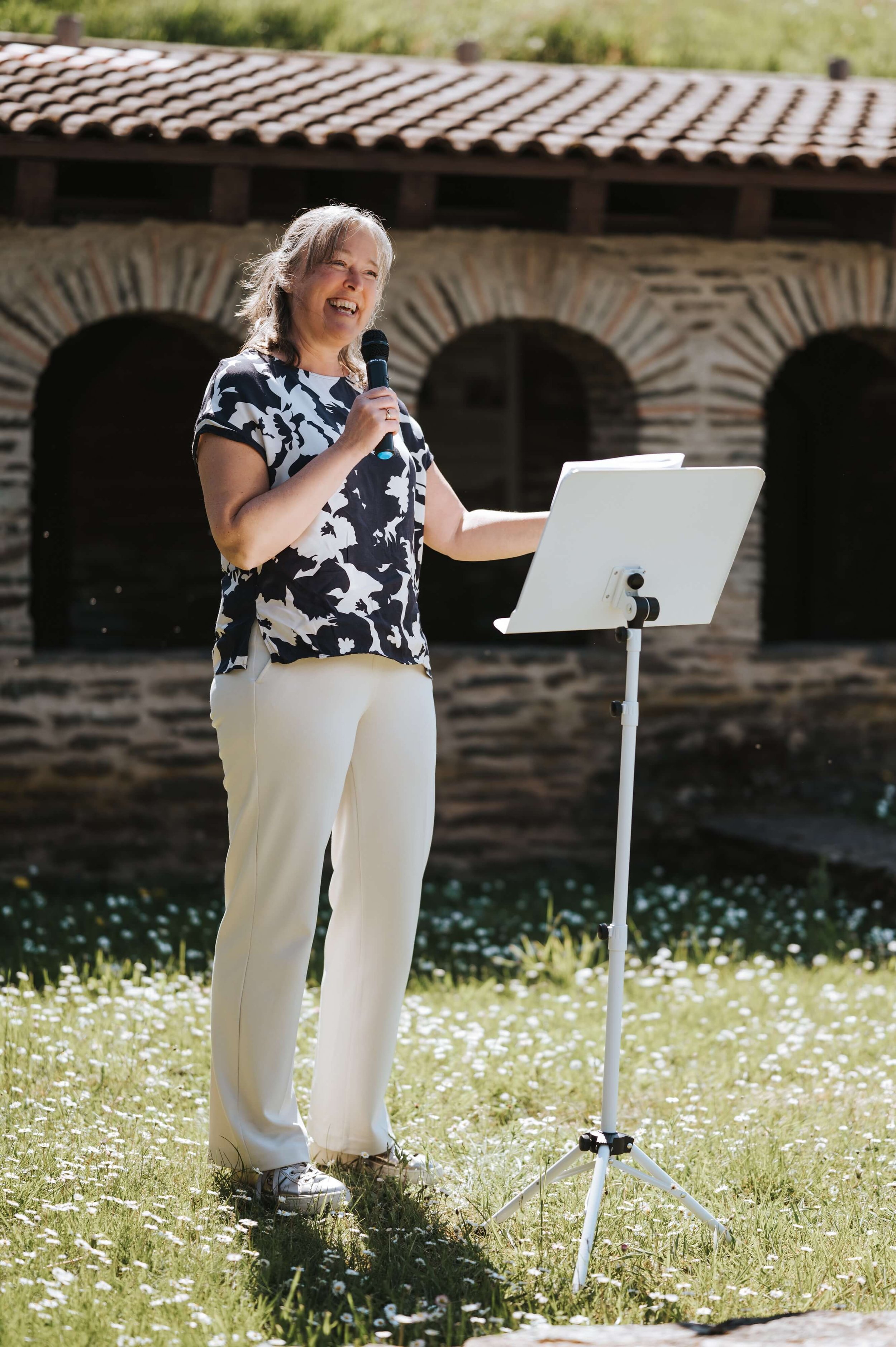 Eine Frau hält einen Mikrofon und steht vor einem Notenständer im Freien, während sie spricht.