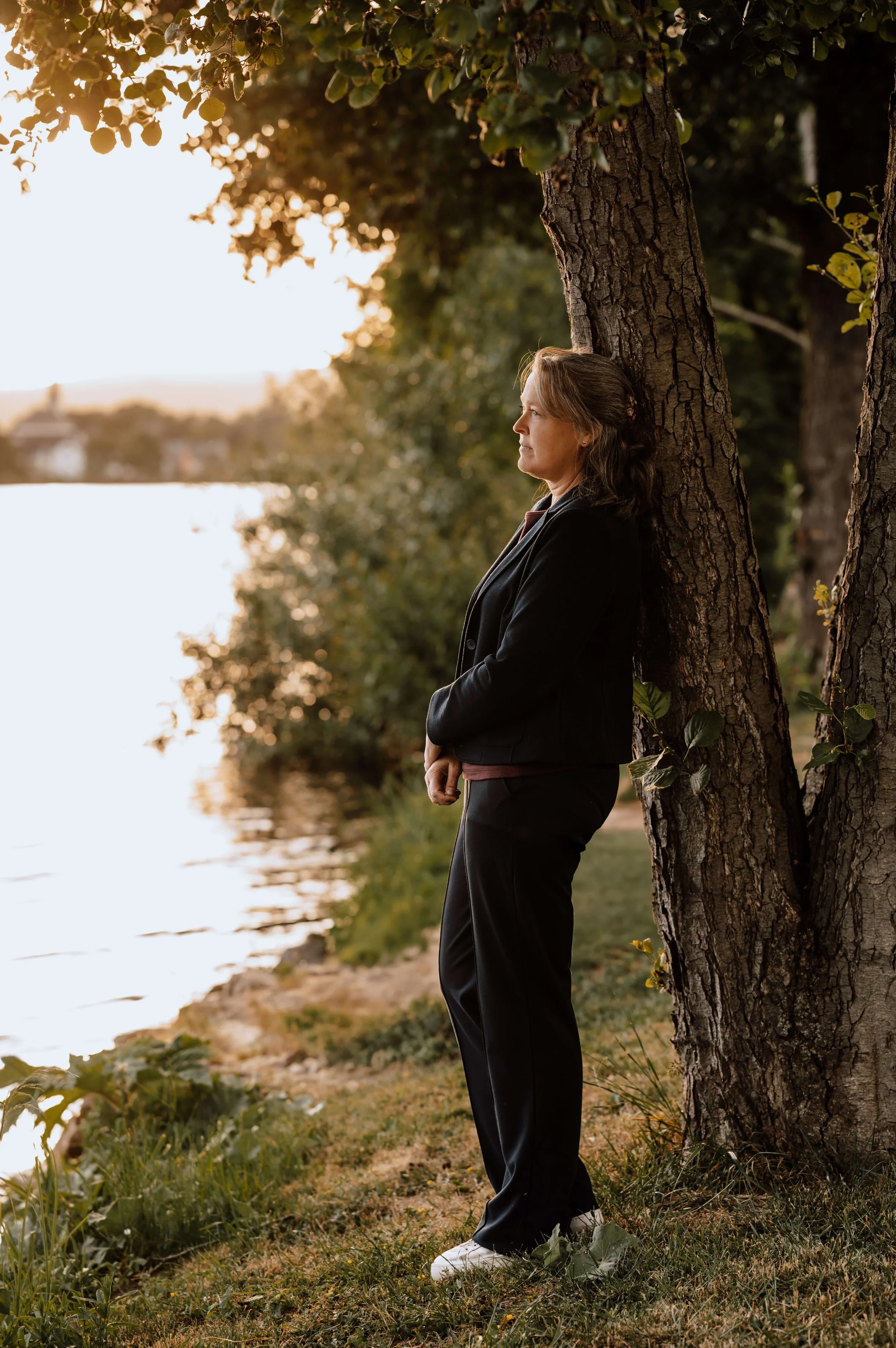 Eine Frau steht an einem Baum am Ufer eines Sees oder Flusses, blickt nach rechts, trägt einen dunklen Anzug und weiße Schuhe, bei Sonnenuntergang, die Umgebung ist grün mit Bäumen und Wasser