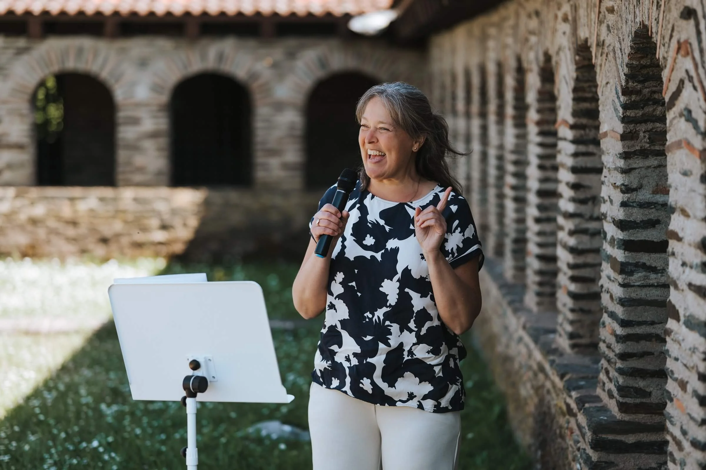 Eine ältere Frau hält ein Mikrofon in der Hand und zeigt mit dem Zeigefinger nach oben, während sie im Freien vor einer alten Backsteinmauer spricht.