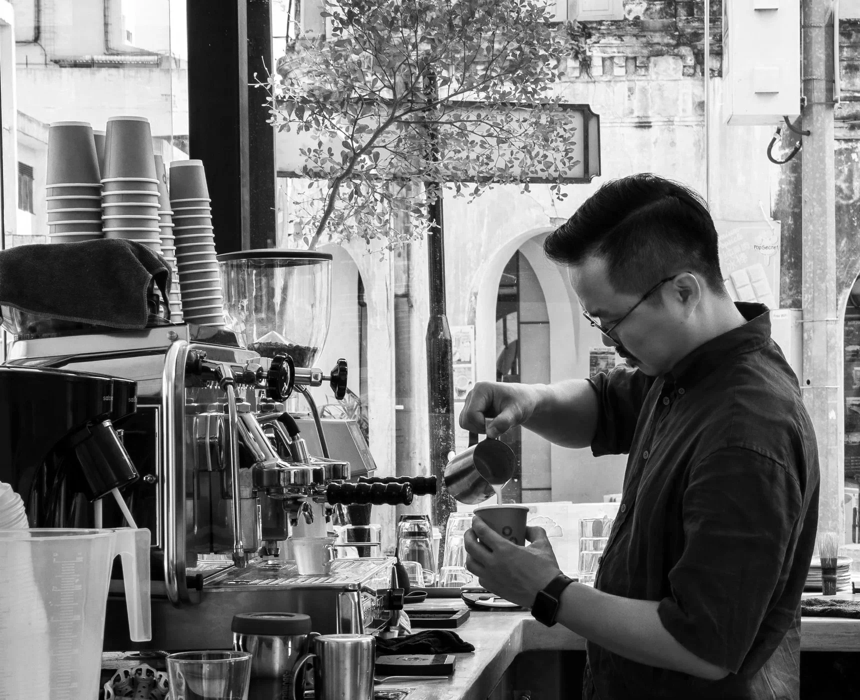 Barista preparing coffee behind a counter in a coffee shop, with a commercial espresso machine and various cups and equipment visible.
