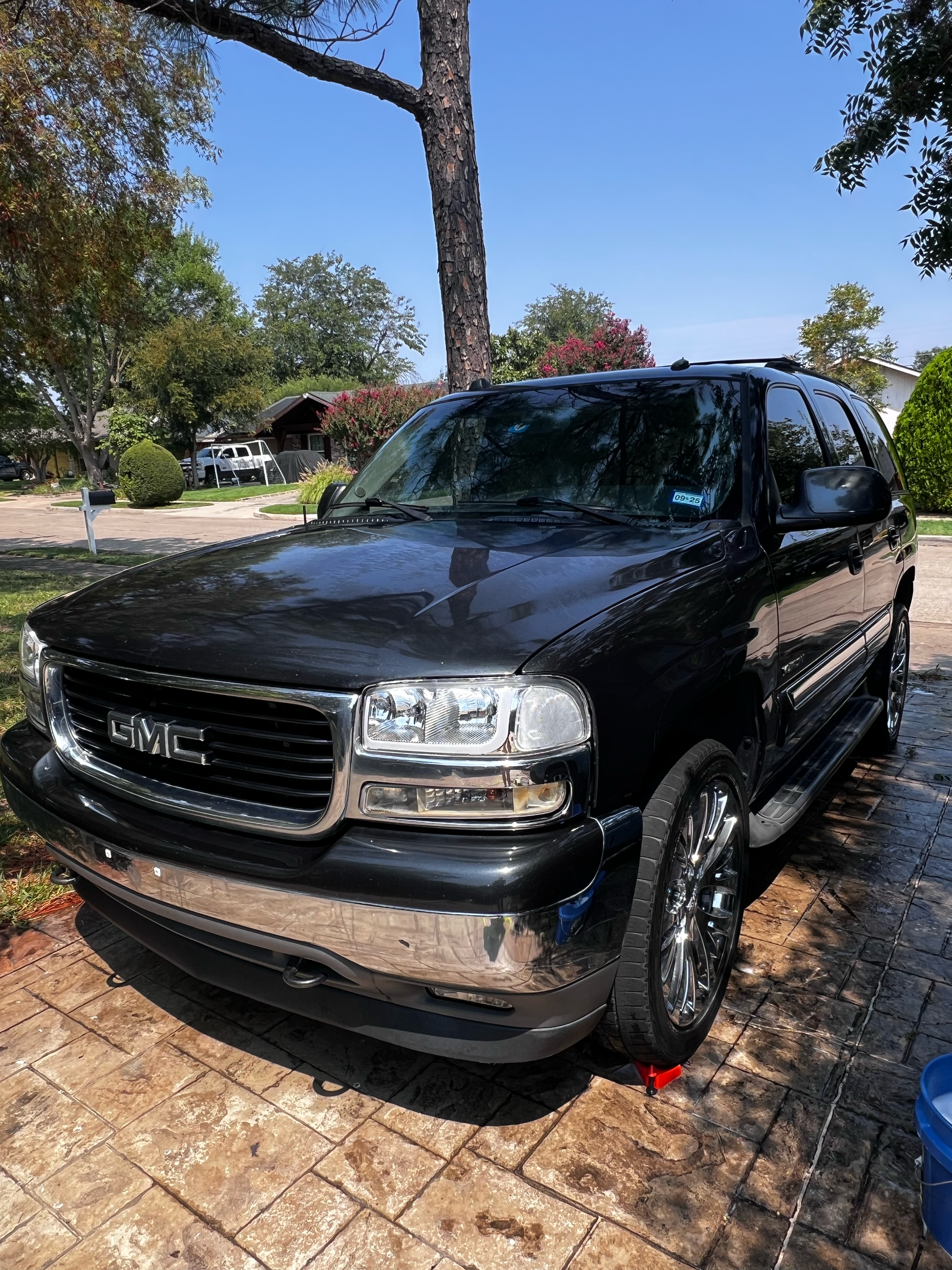 Black GMC SUV parked on a brick driveway under a tree in a suburban neighborhood with houses and greenery in the background on a sunny day.