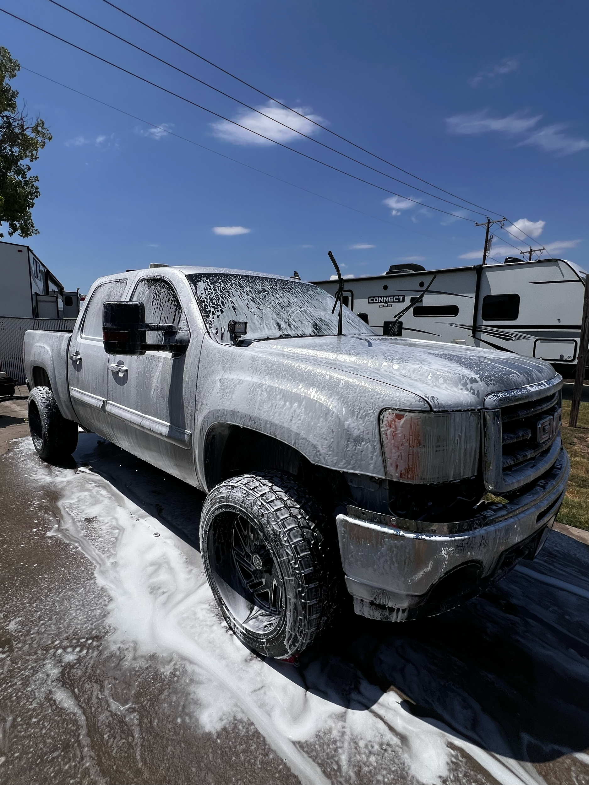 Silver pickup truck covered in soap suds during washing, with foam on the ground underneath, under a blue sky with some clouds, and other vehicles and trailers in the background.