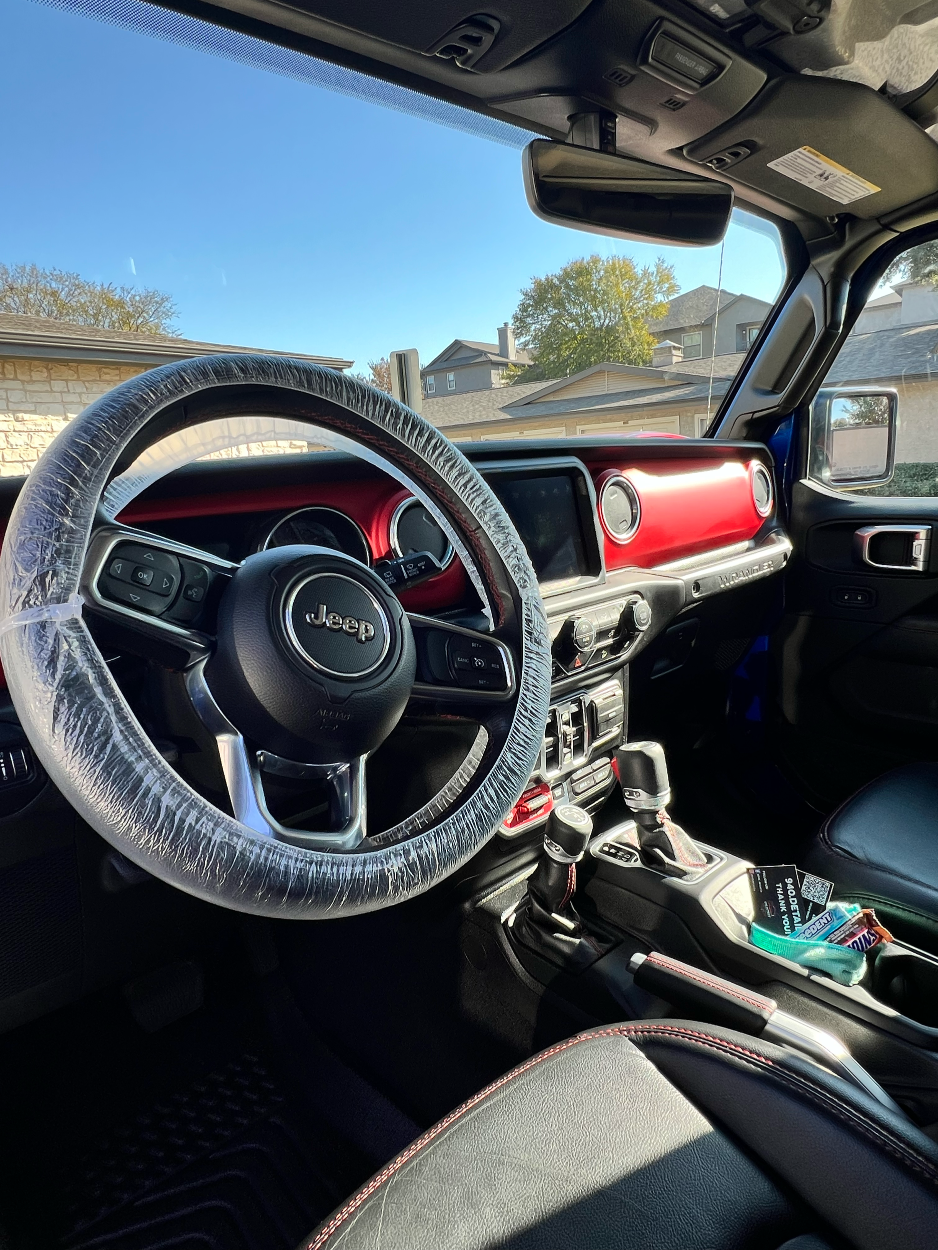 The interior of a Jeep vehicle featuring a steering wheel with a plastic cover, red dashboard accents, gear shifters, and some snacks in the front seat cup holder, with a view of houses past the windshield.