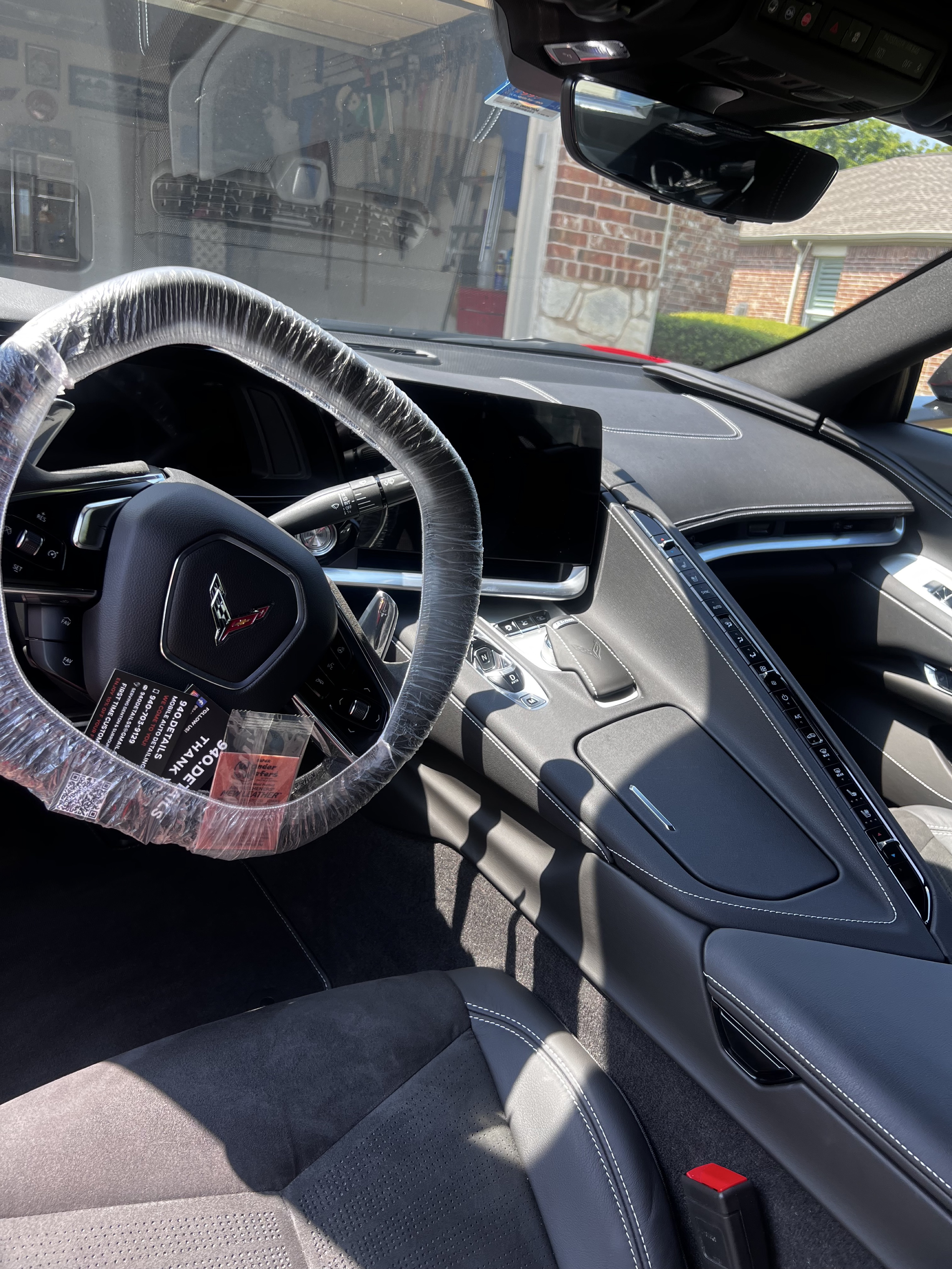 Interior of a Chevrolet Corvette with a steering wheel wrapped in protective plastic and a digital dashboard. Visible are the center console, a touchscreen display, and the passenger seat. The interior features black and gray materials with white sti