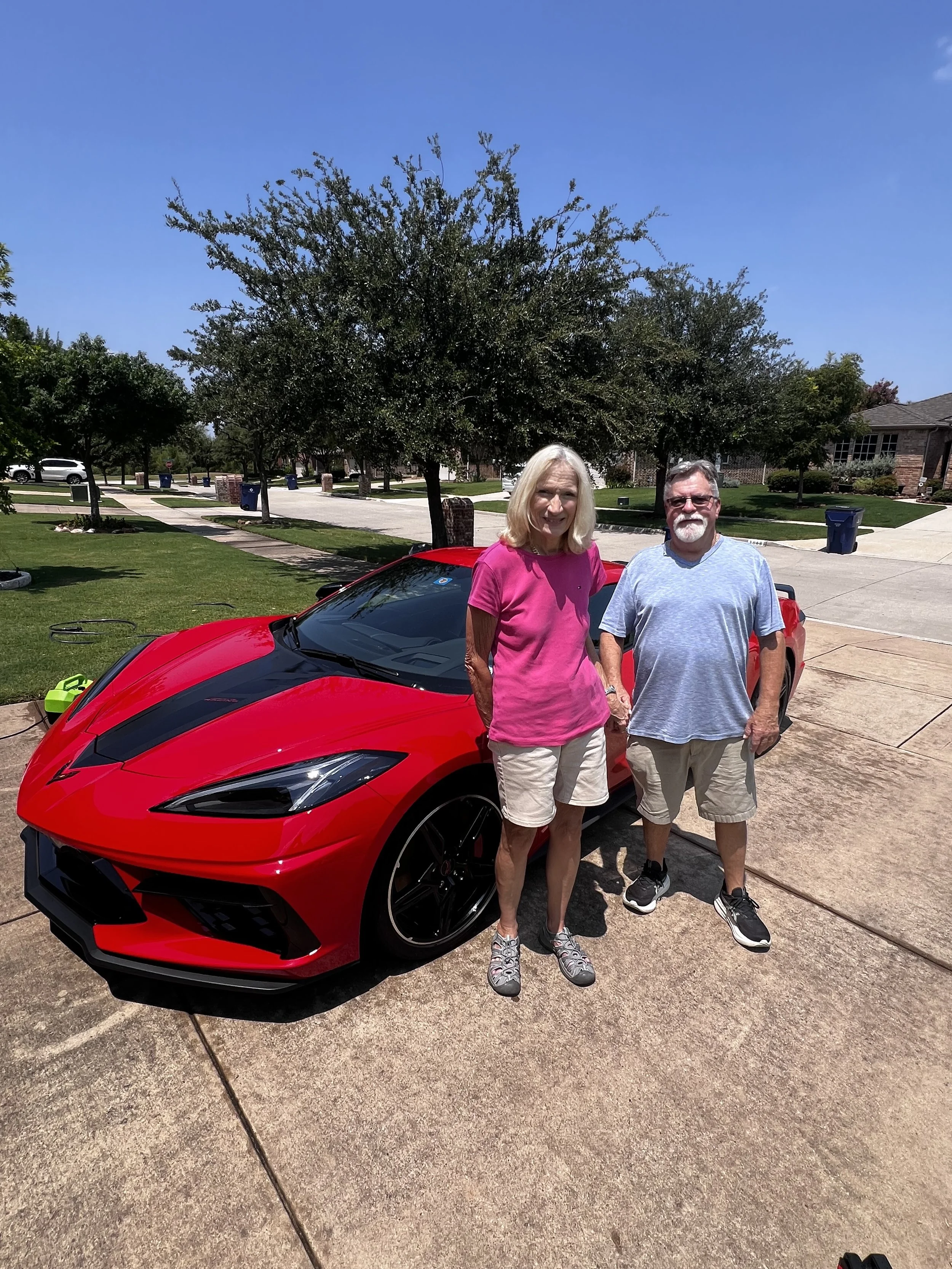 A woman and a man standing outside on a driveway next to a red sports car under a clear blue sky.
