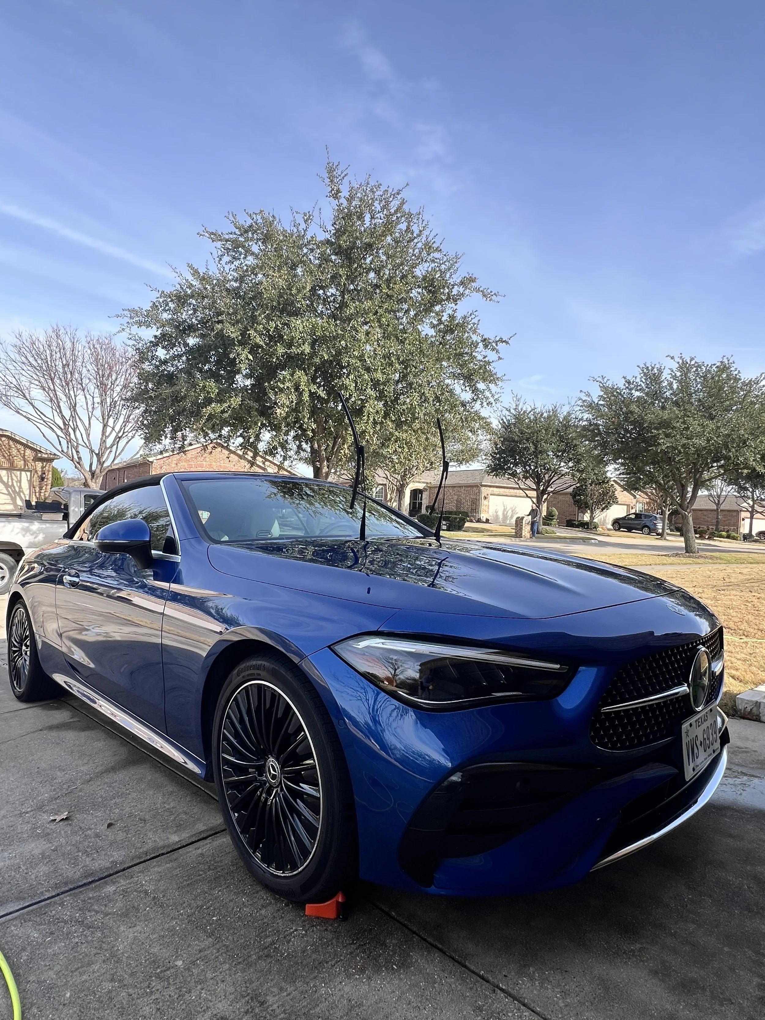 A blue convertible car parked on a driveway with a red wheel lock. There are trees and houses in the background under a clear blue sky.