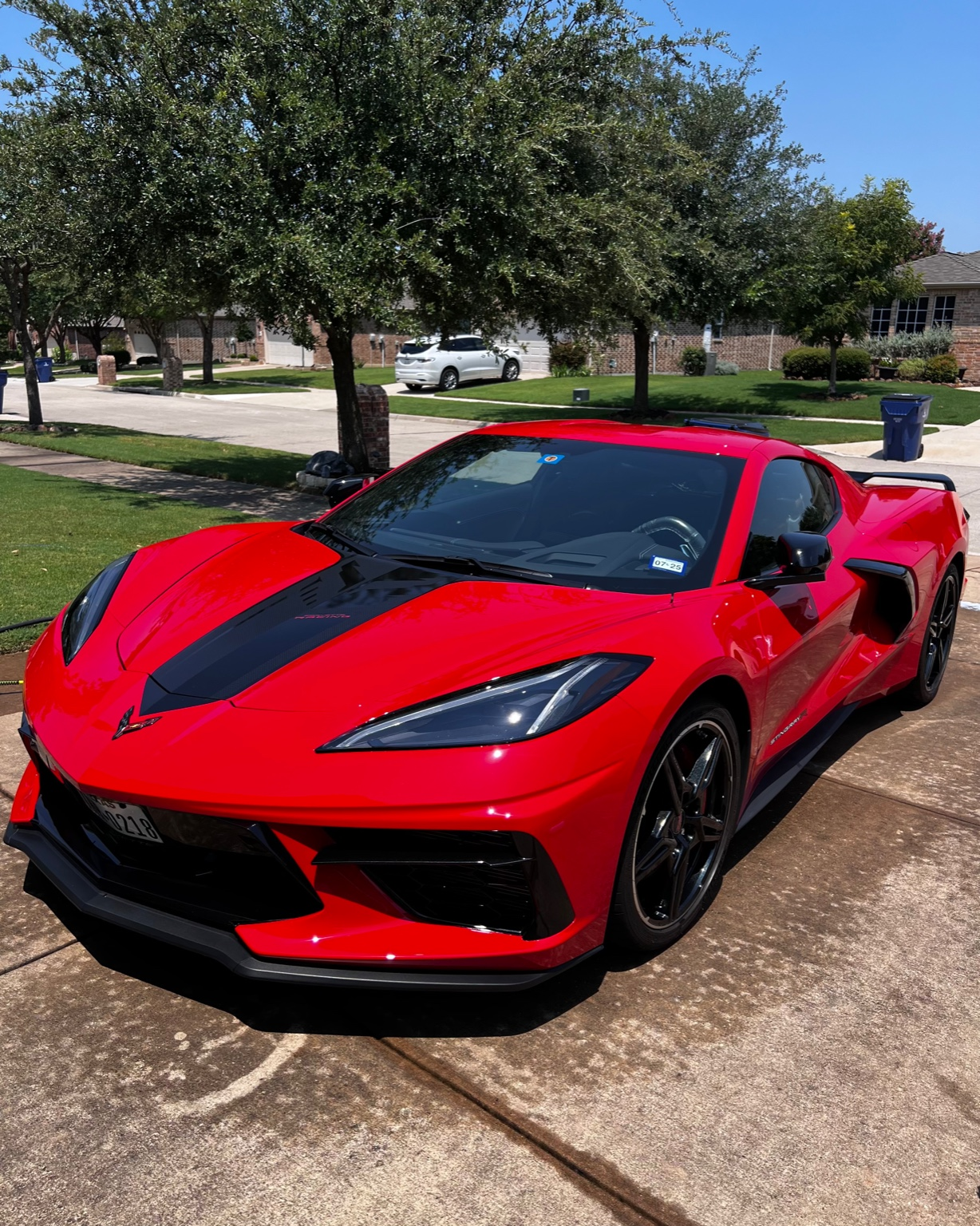 Red sports car with black racing stripe parked on driveway in front of suburban houses with green lawns and trees.