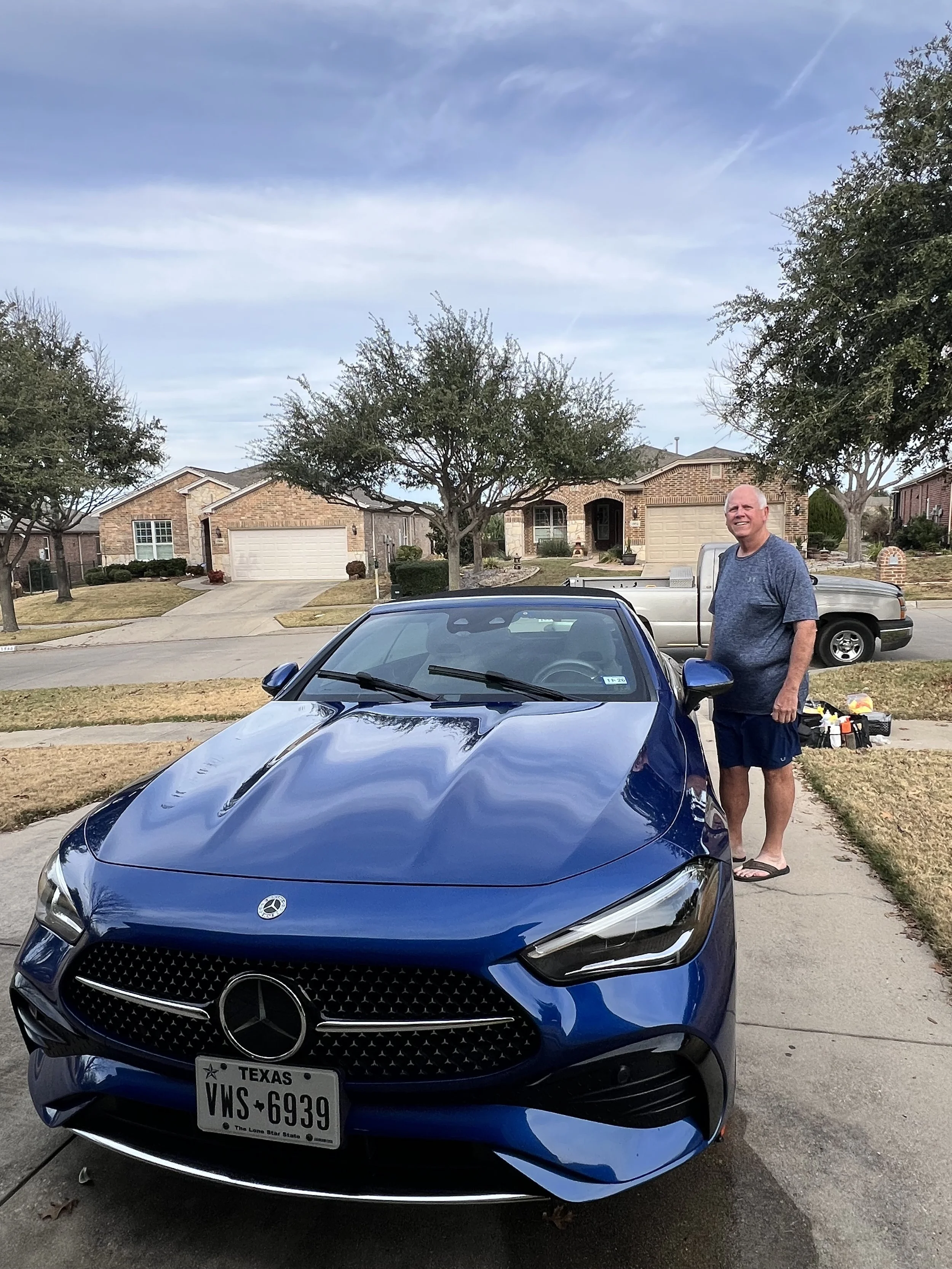 A man standing next to a blue Mercedes-Benz sports car parked in a suburban neighborhood driveway. There are houses, trees, and another parked vehicle in the background.
