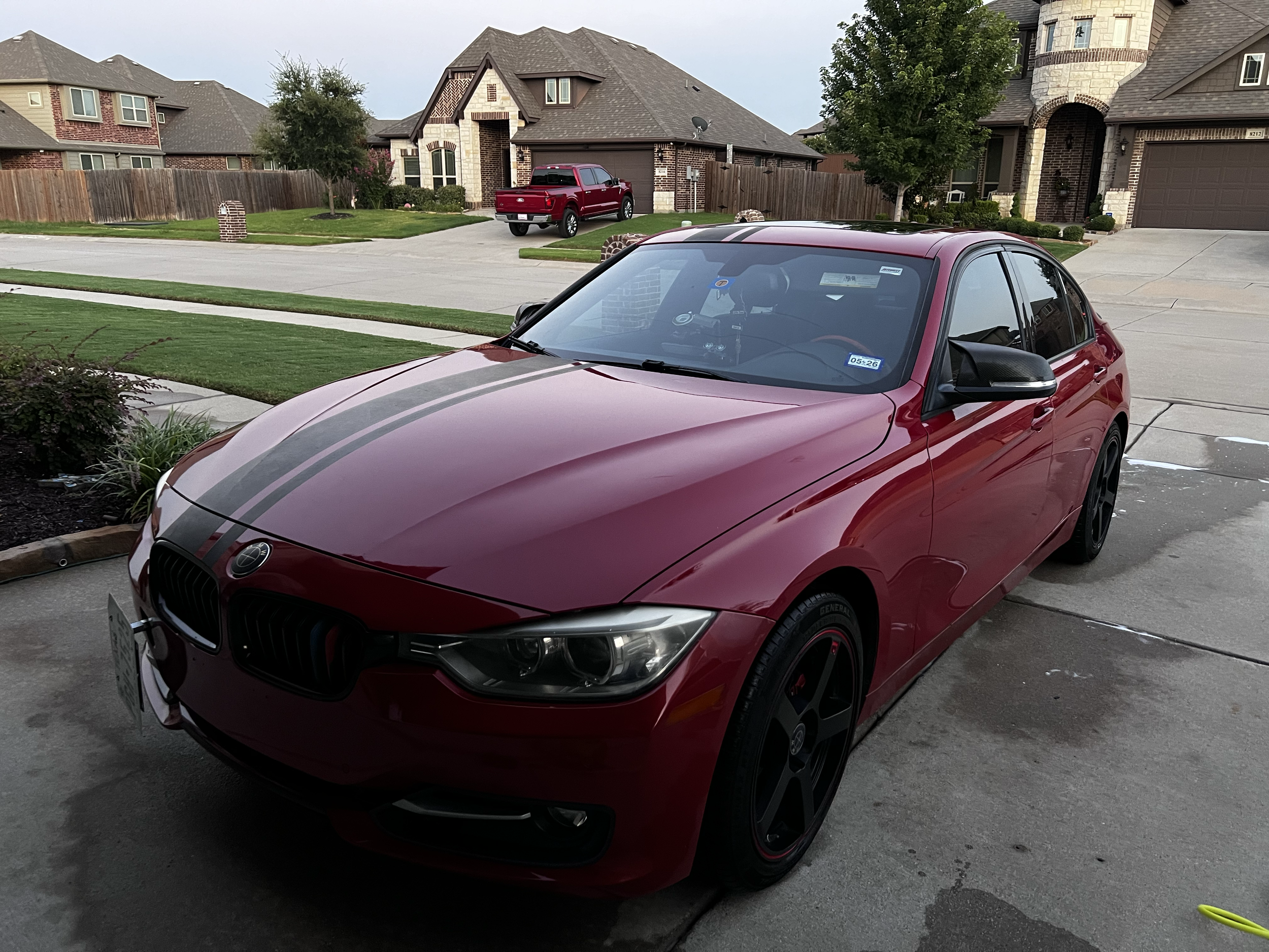 A red sports car with black racing stripes parked in a residential driveway with houses and a street in the background.