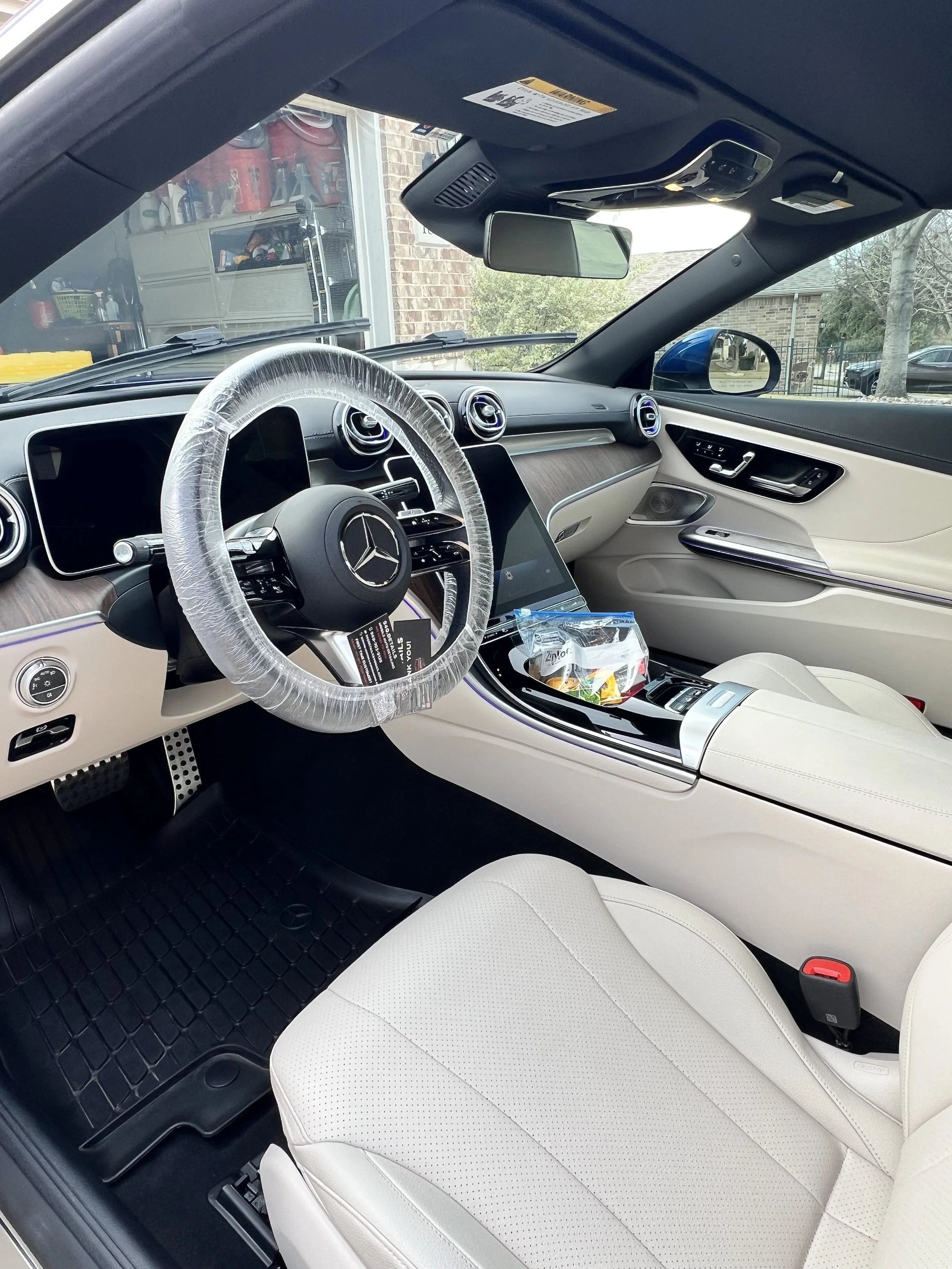 Interior of a Mercedes-Benz vehicle with white leather seats, wood trim, and a dashboard featuring a large touchscreen display. The steering wheel is covered with plastic wrap.