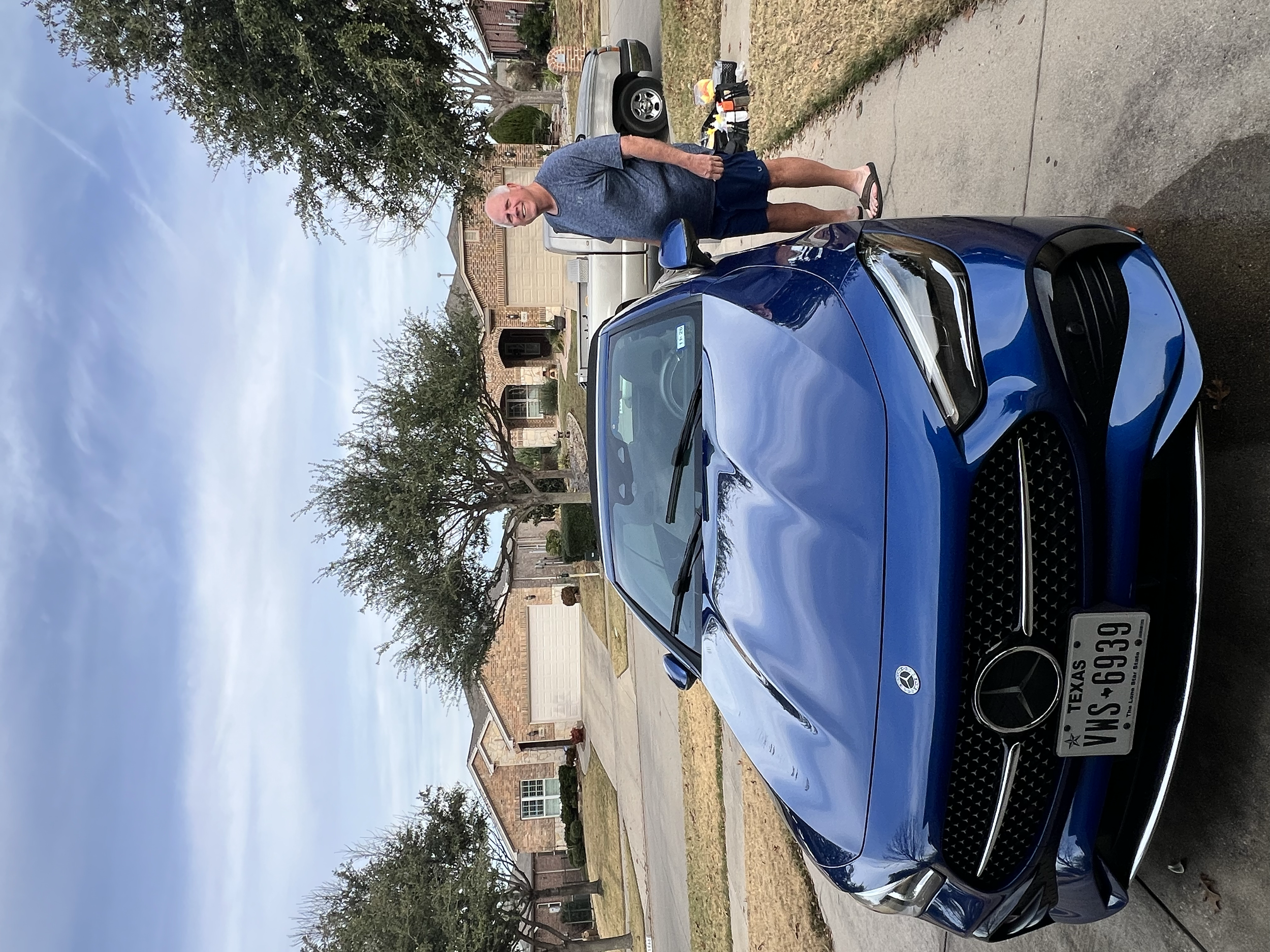 A bald man wearing a gray T-shirt, black shorts, and flip-flops standing next to a blue Mercedes-Benz parked in a residential driveway with brick houses and trees in the background.