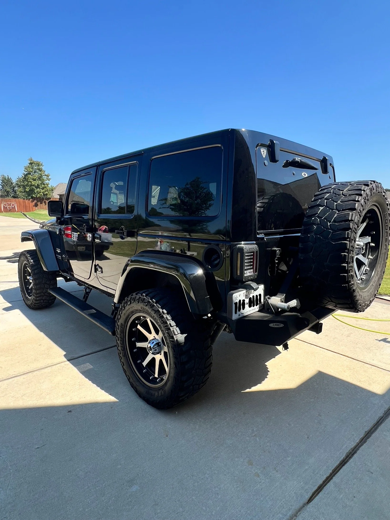 Black Jeep Wrangler with off-road tires parked on a driveway under clear blue sky.