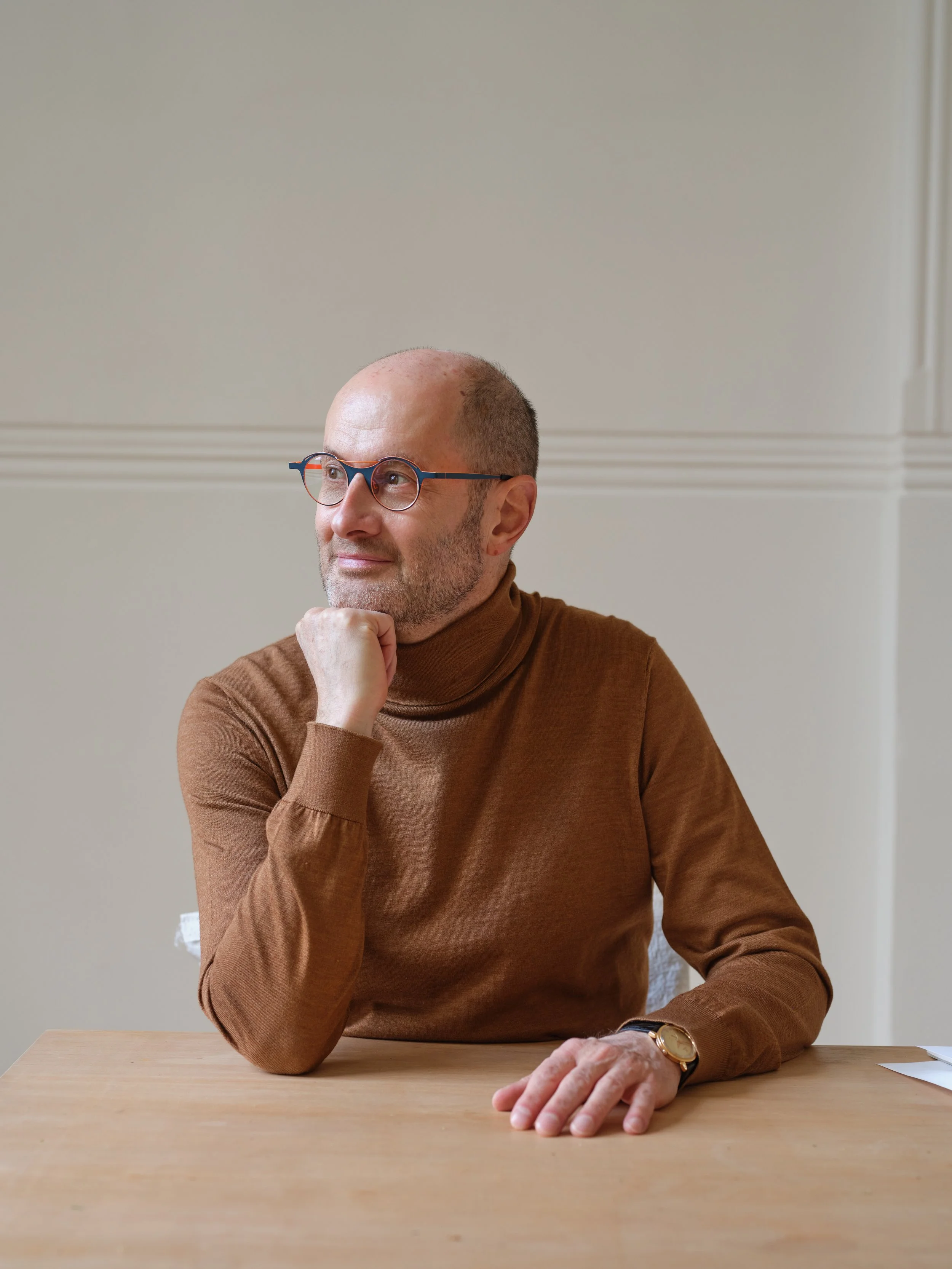 A man in a brown turtleneck shirt sitting at a wooden table, wearing glasses and a wristwatch, with his chin resting on his hand, looking thoughtfully to the side in a neutral-colored room.