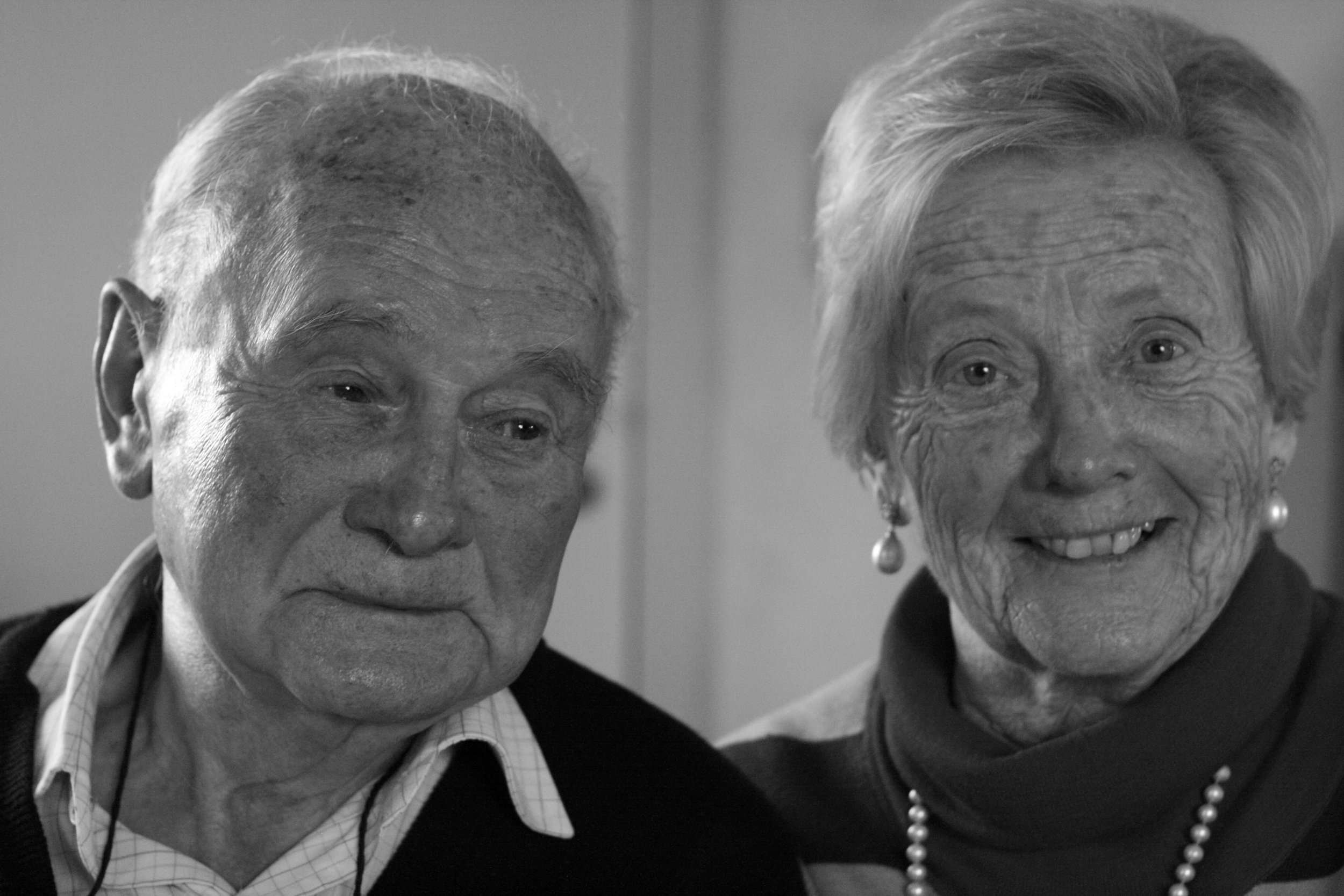 Black and white photo of an elderly man and woman sitting close to each other, both smiling softly. The woman is wearing pearl earrings and a pearl necklace.