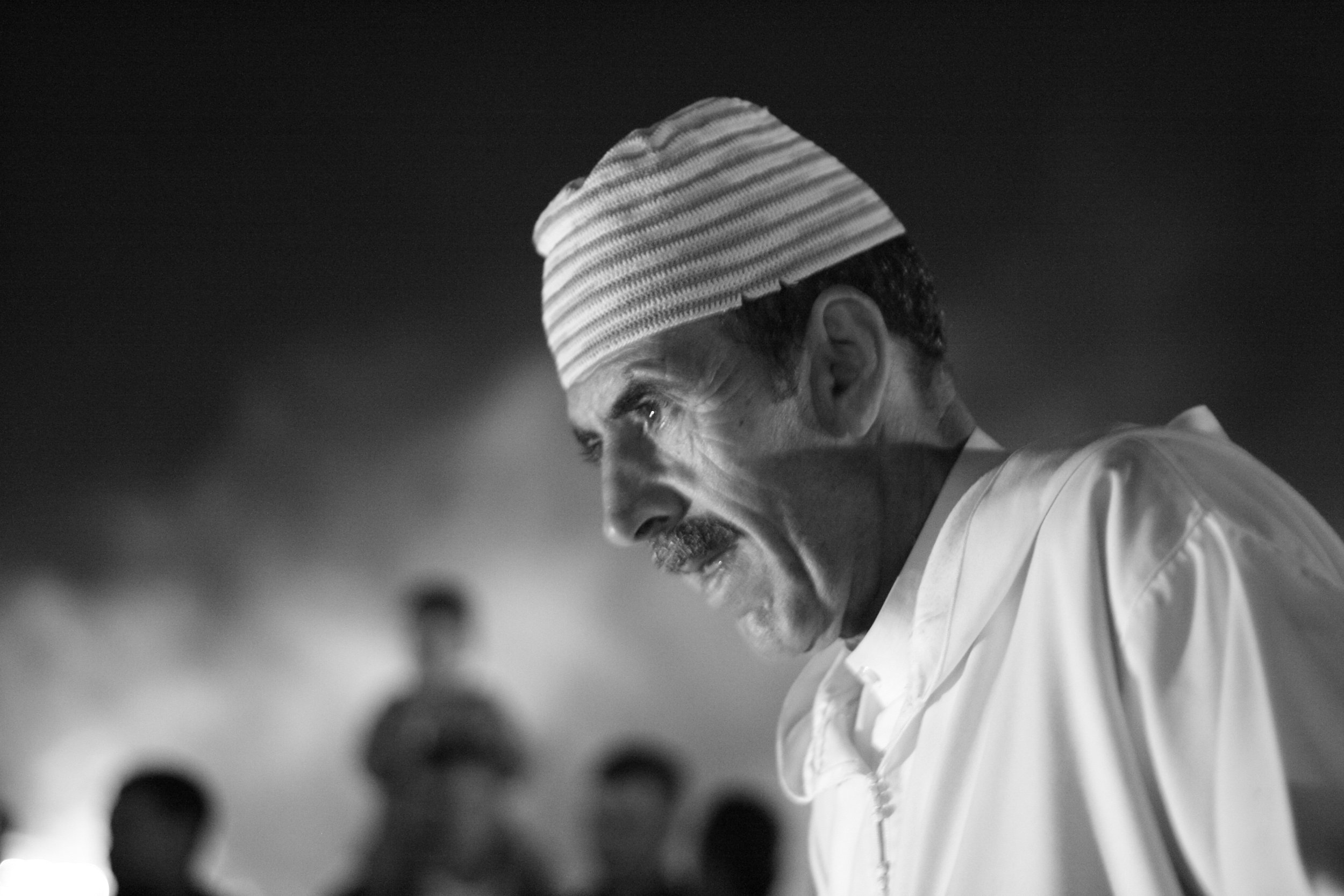 A black-and-white photograph of an older man wearing a traditional cap, with a serious expression, looking downward with a solemn or intense demeanor. Blurred figures are in the background.