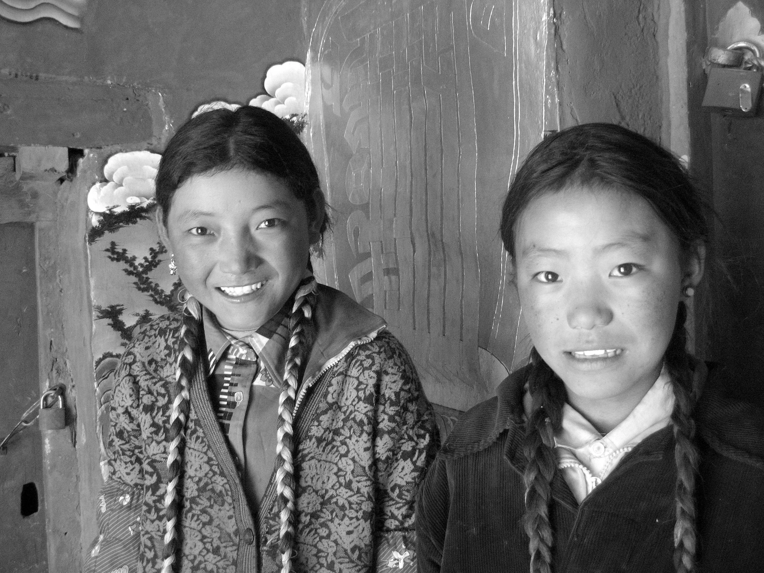 Two young girls with braided hair smiling and standing inside a room with a decorated wall behind them.