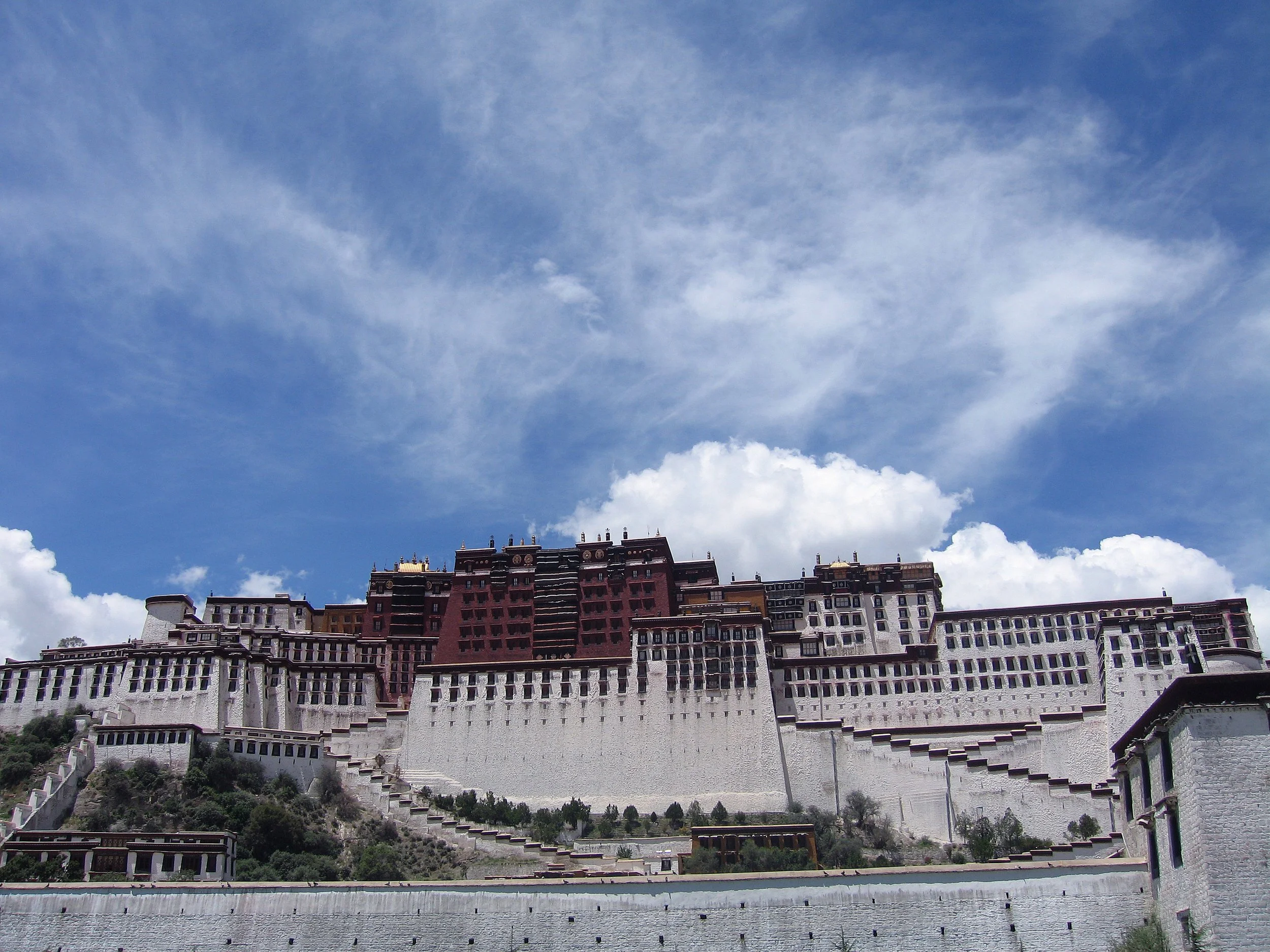 A large palace or monastery complex with multiple levels and white walls, situated on a hillside under a partly cloudy blue sky.