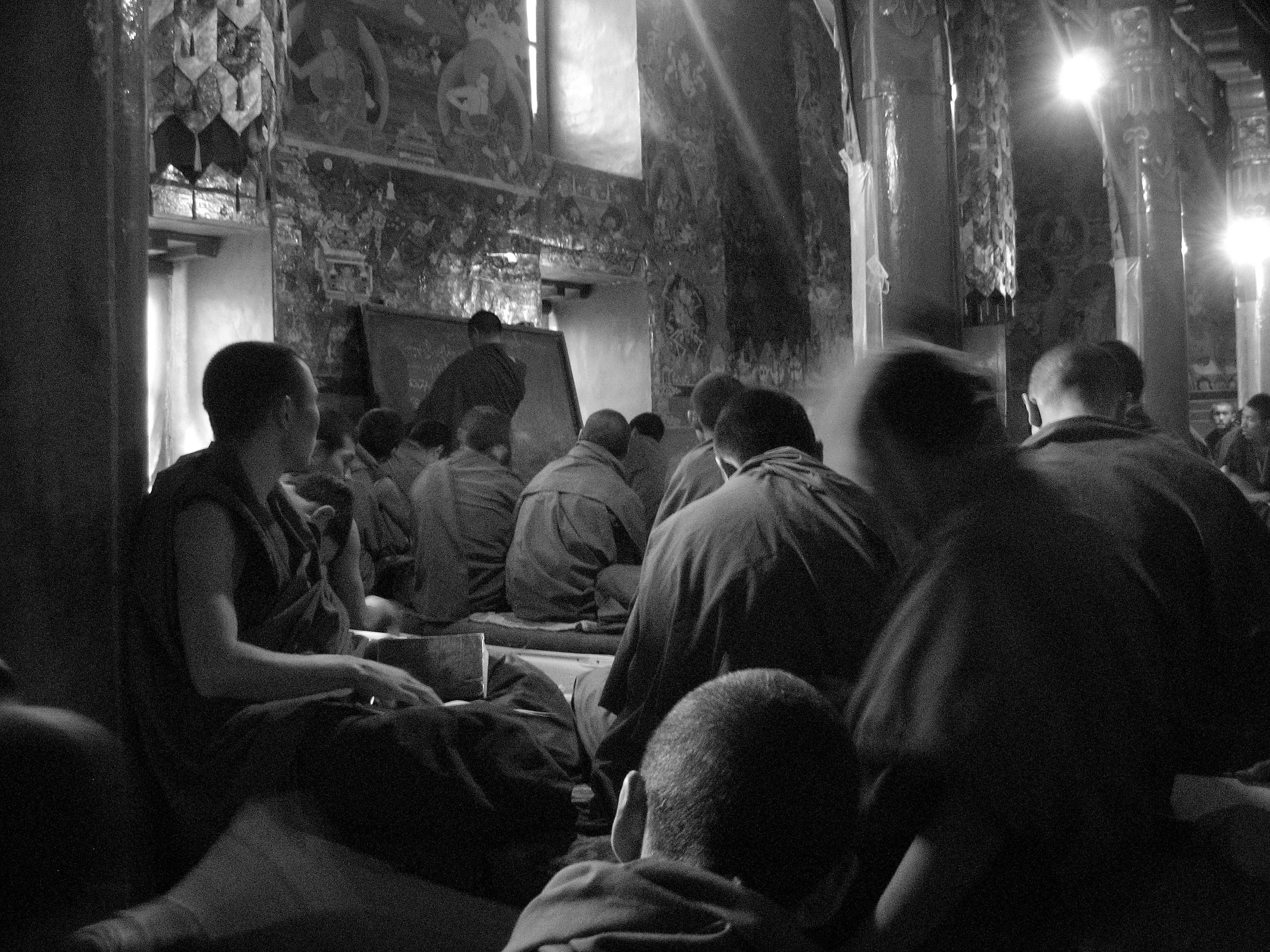 Group of monks or religious individuals sitting on the floor inside a temple or monastery, facing a chalkboard where a person is writing or presenting. The interior features ornate wall decorations and pillars with intricate designs, and light stream