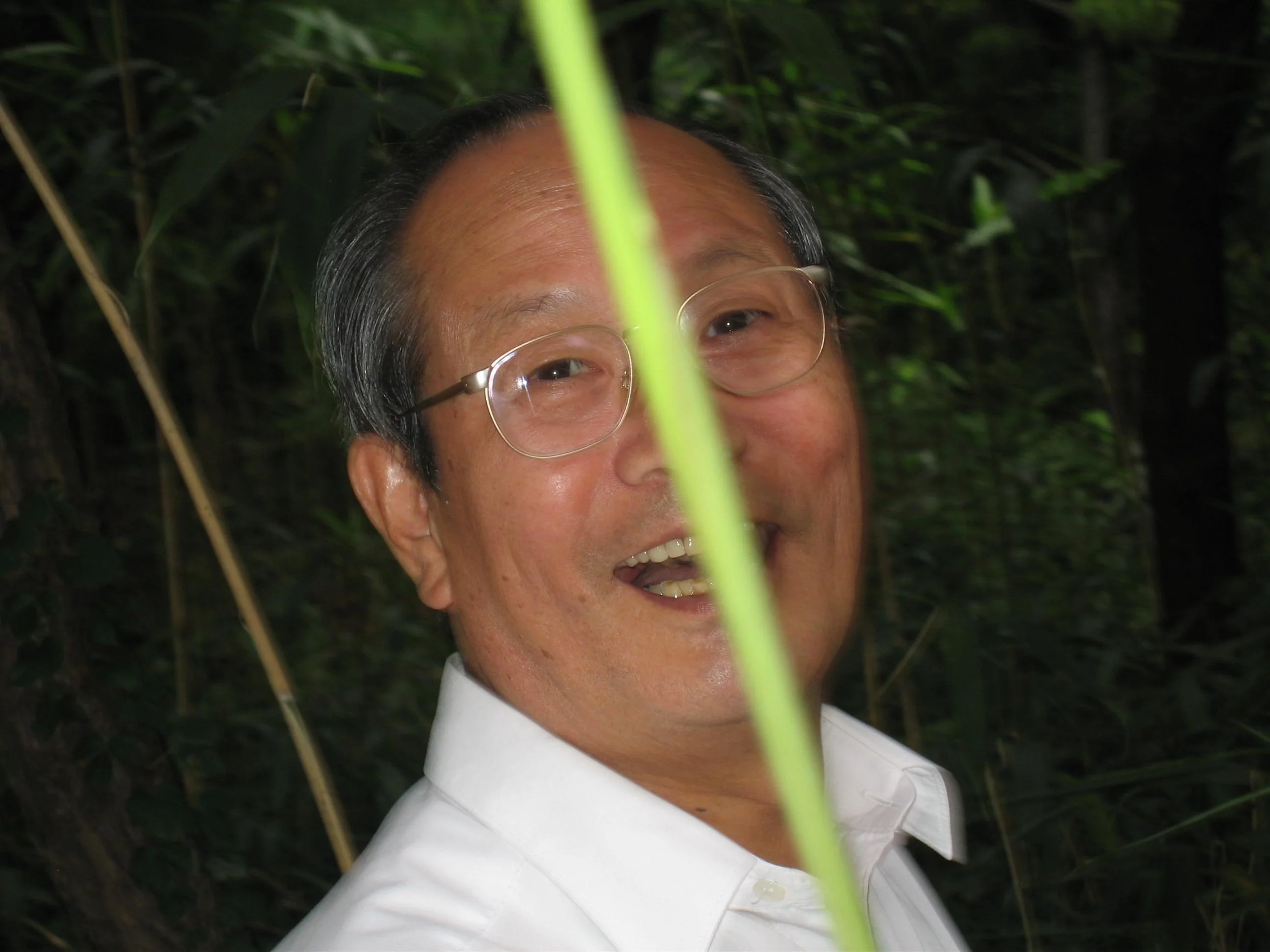 A smiling man with glasses, wearing a white shirt, partially obscured by a green plant in a dense forest or jungle.