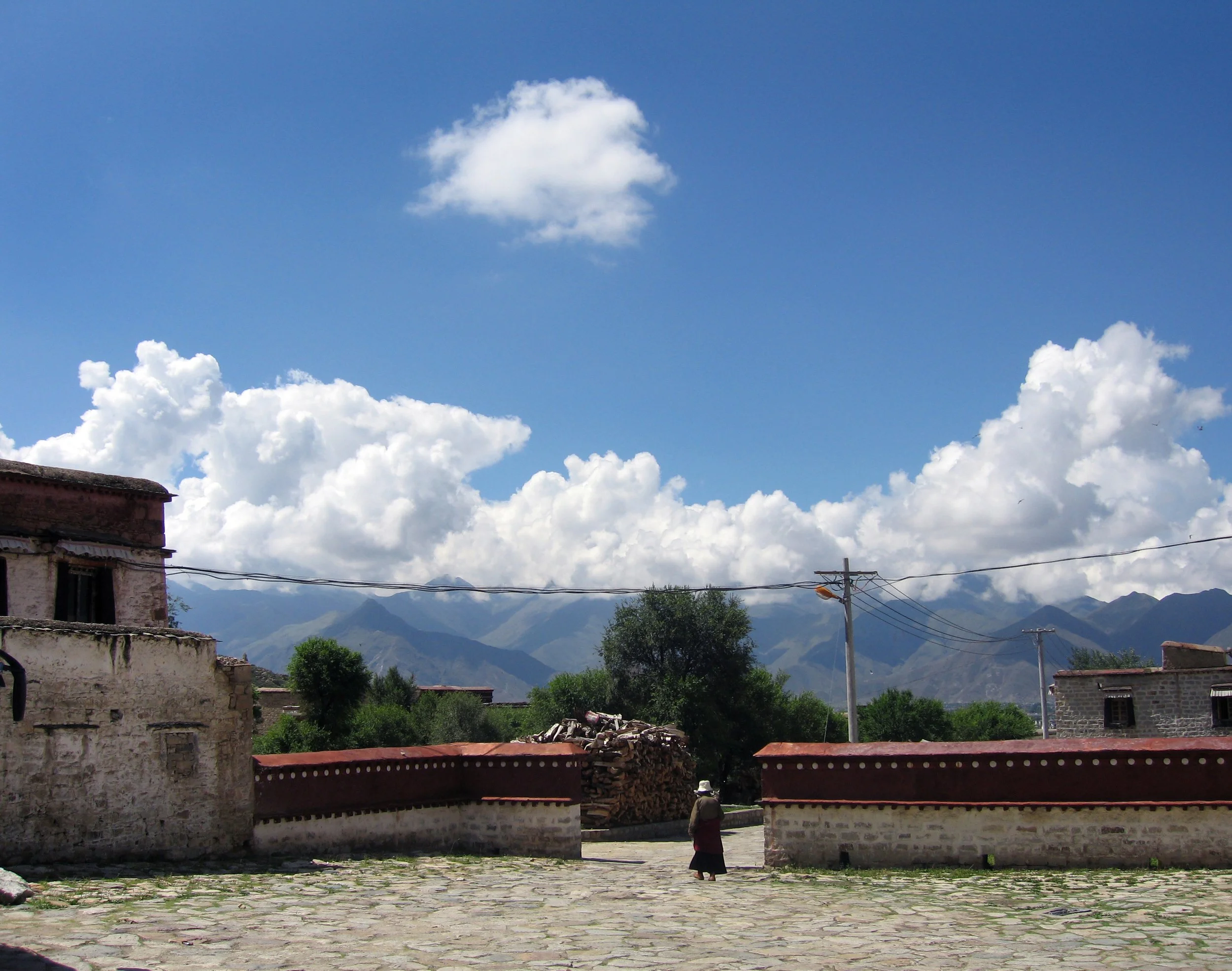 A rural village scene with stone buildings, a red brick wall, a woman wearing traditional clothing and a hat, trees, and mountains under a partly cloudy blue sky.