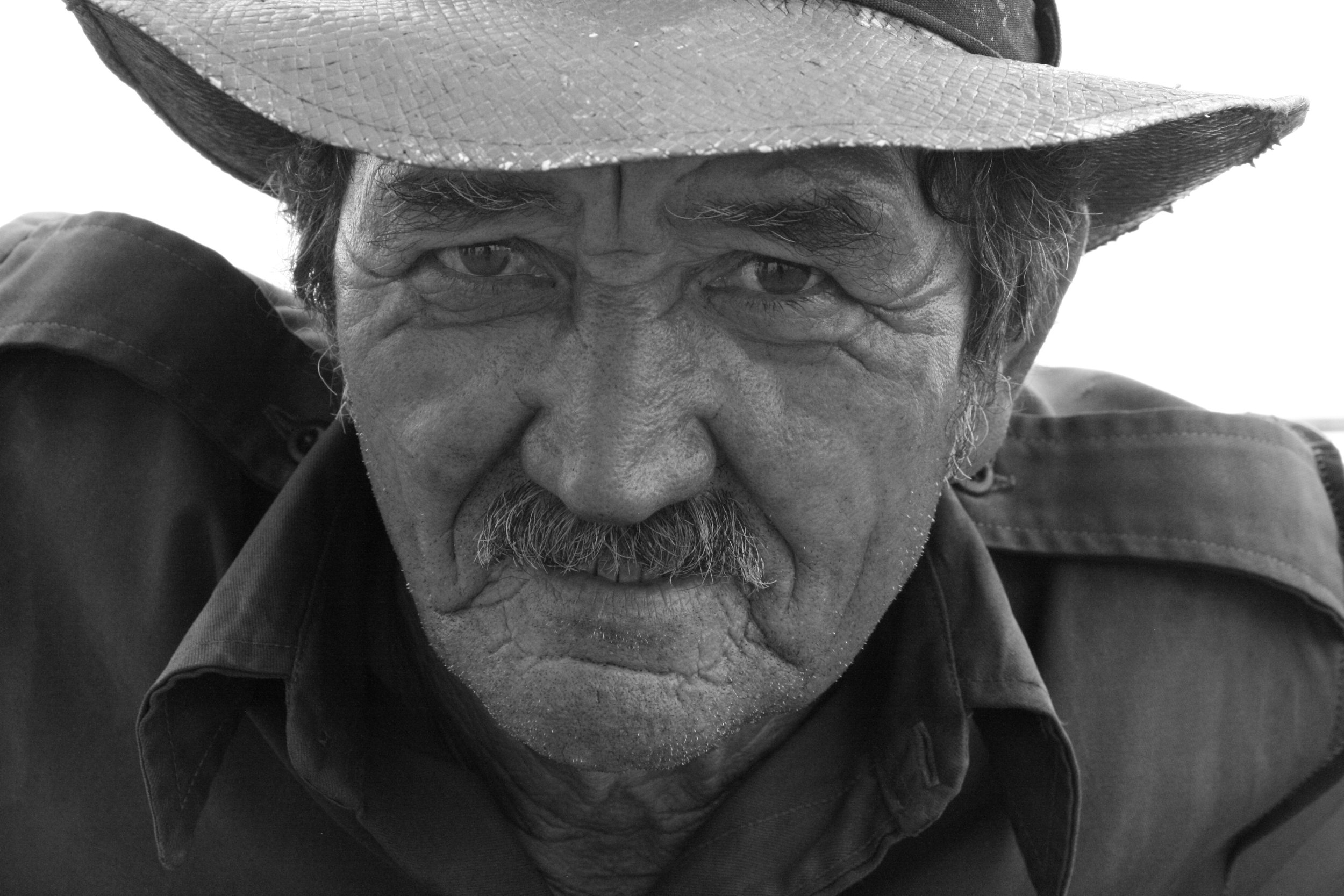 Close-up of an elderly man with weathered skin, wearing a cowboy hat and a dark shirt, gazing intently at the camera.
