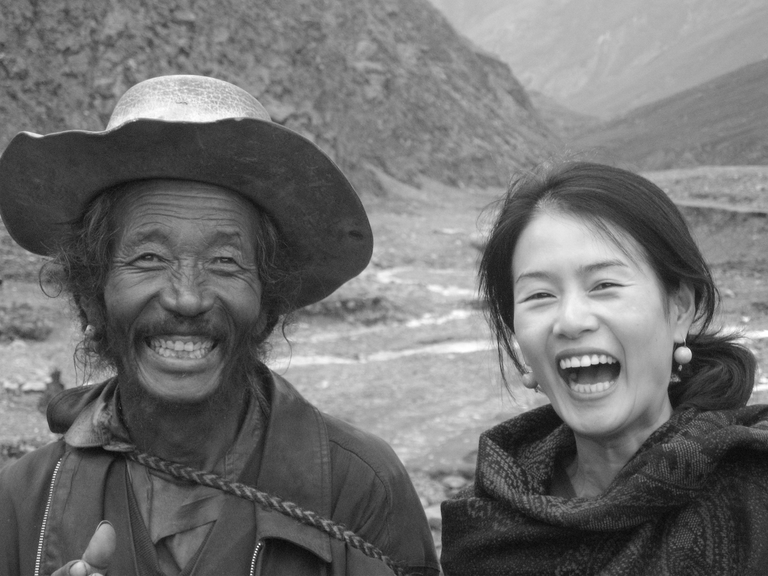 A black and white photo of a smiling elderly man wearing a wide-brimmed hat and a woman with long hair laughing, outdoors in a mountainous area.