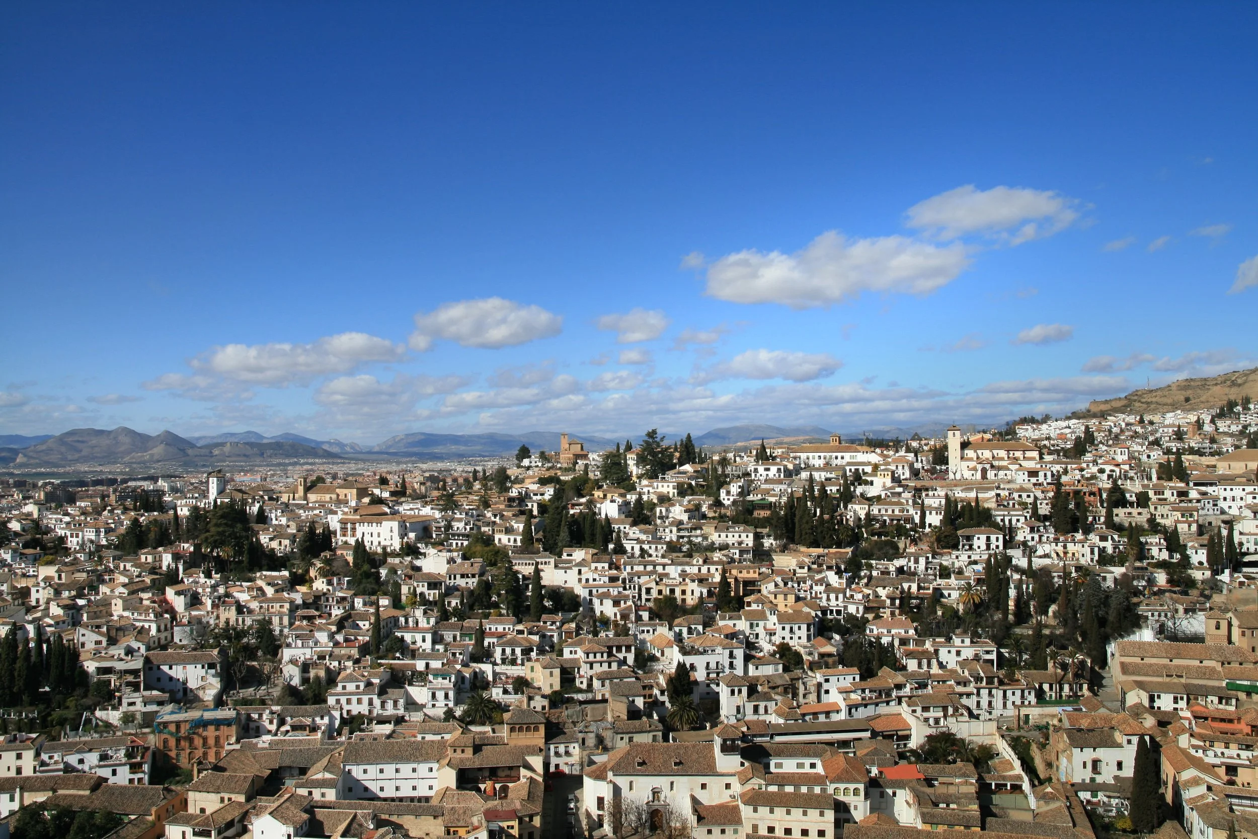 A panoramic view of a city with white buildings, terracotta rooftops, trees, and hills in the background under a blue sky with scattered clouds.