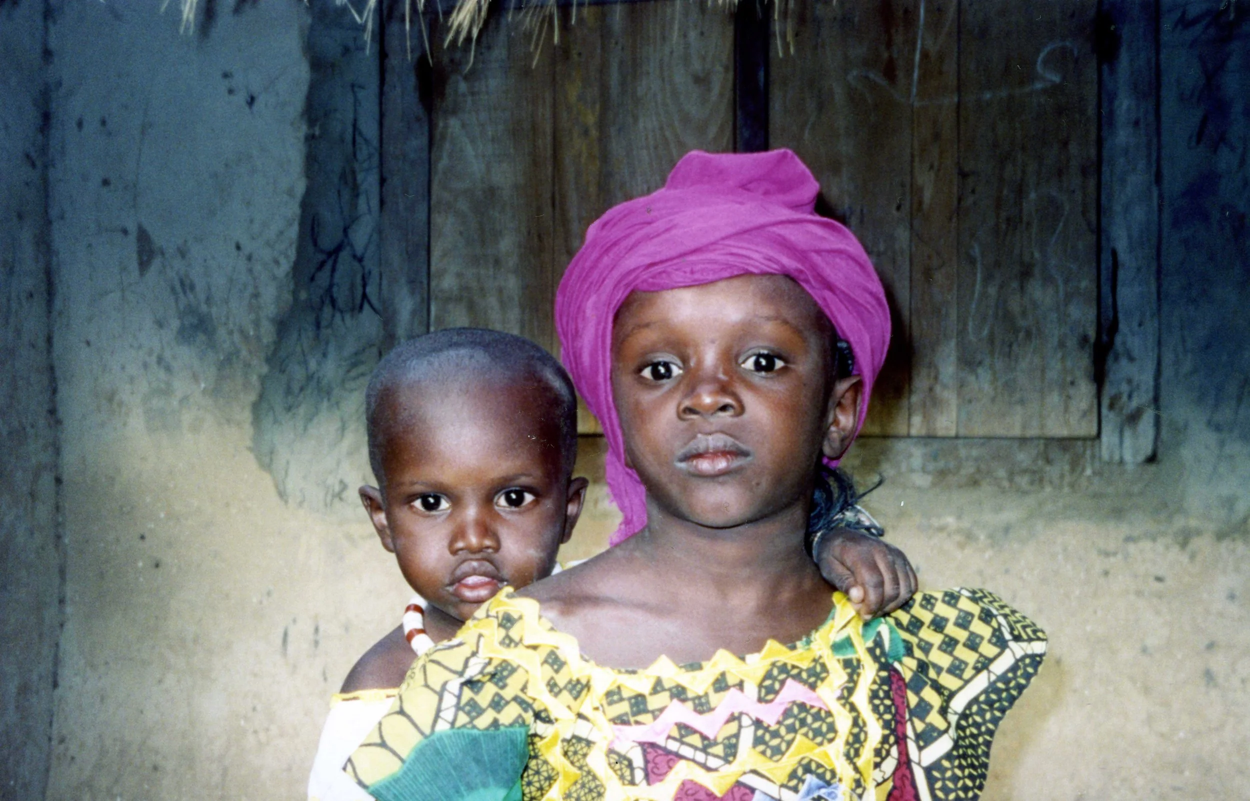Two young children, a girl in a colorful patterned dress and a purple headscarf, and a boy with a beaded necklace, standing inside a rustic building with wooden walls.