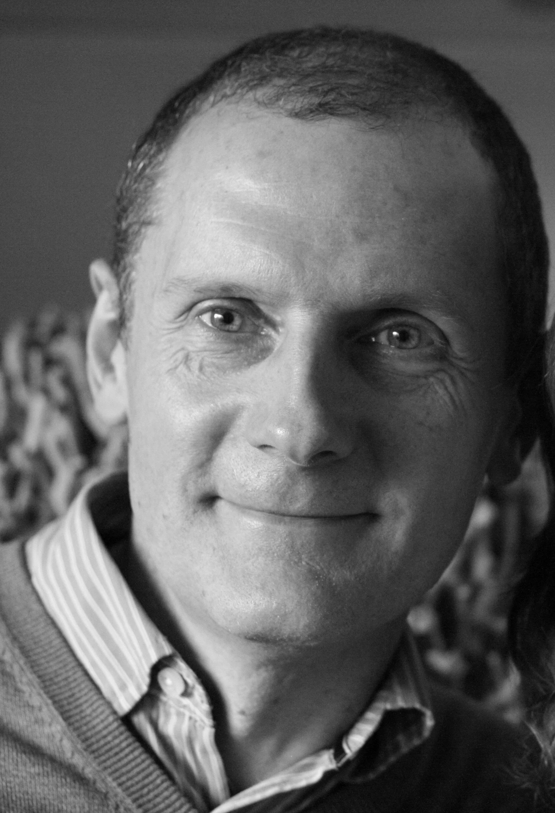A black and white close-up portrait of a man with short hair, smiling subtly, wearing a striped collared shirt and a sweater.