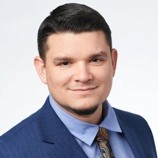 Hombre con barba y cabello rizado, gafas transparentes, usando un suéter negro con camiseta blanca debajo, en un ambiente interior con planta en el fondo