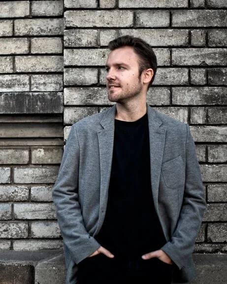 Joven sonriendo con cabello corto y barba, en un entorno al aire libre con cielo nublado y personas en el fondo.