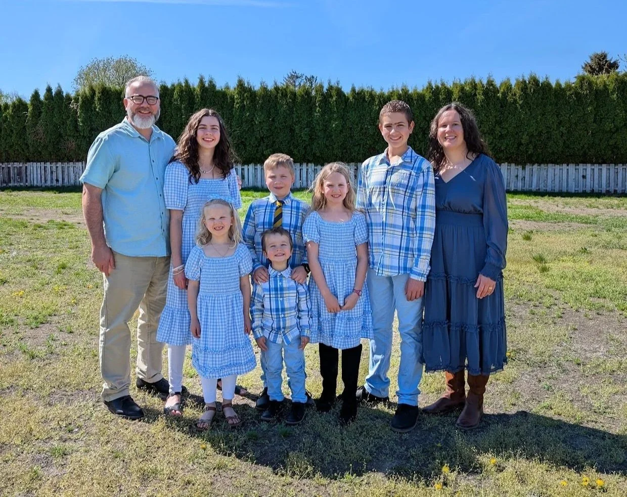A family of ten standing outdoors on a sunny day, dressed in blue and white clothing, smiling with a green hedge and blue sky in the background.