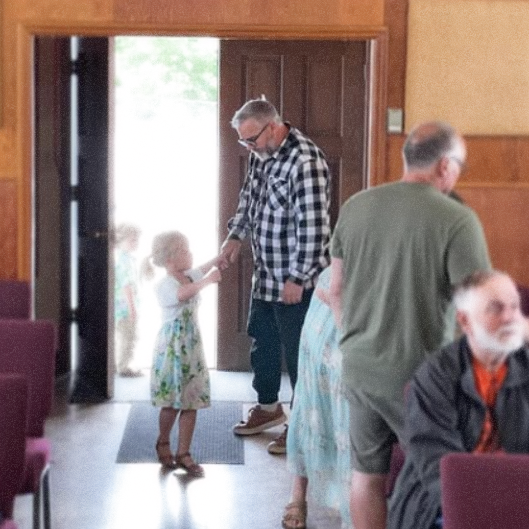 A man wearing glasses and a black-and-white plaid shirt is holding hands with a young girl in a floral dress, standing near the open doorway of a room. Several other people are present in the room, some seated and some standing, with one person partially visible in the foreground.