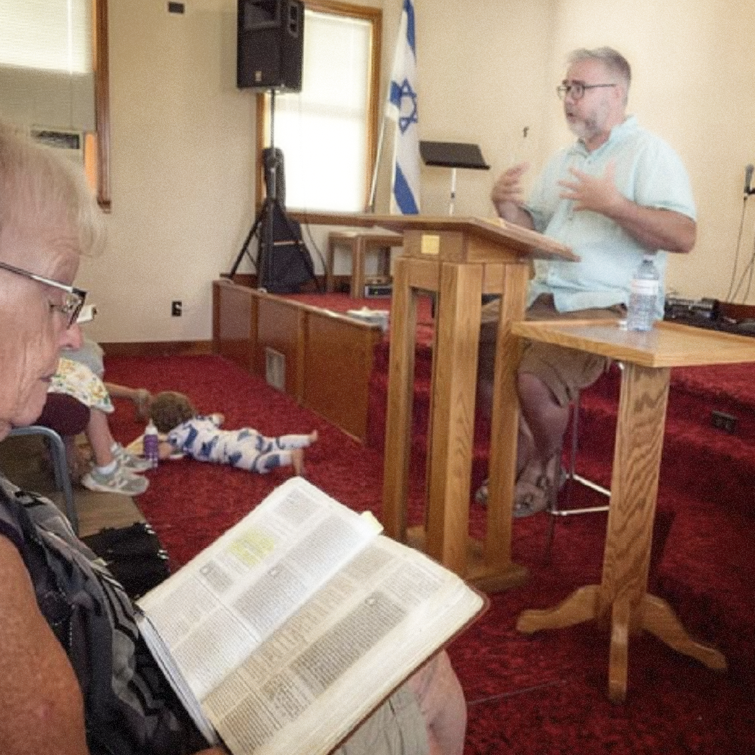 A man giving a speech at a podium in a room decorated with an Israeli flag, with a few children sitting on the carpet and an older woman reading a Bible in the foreground.