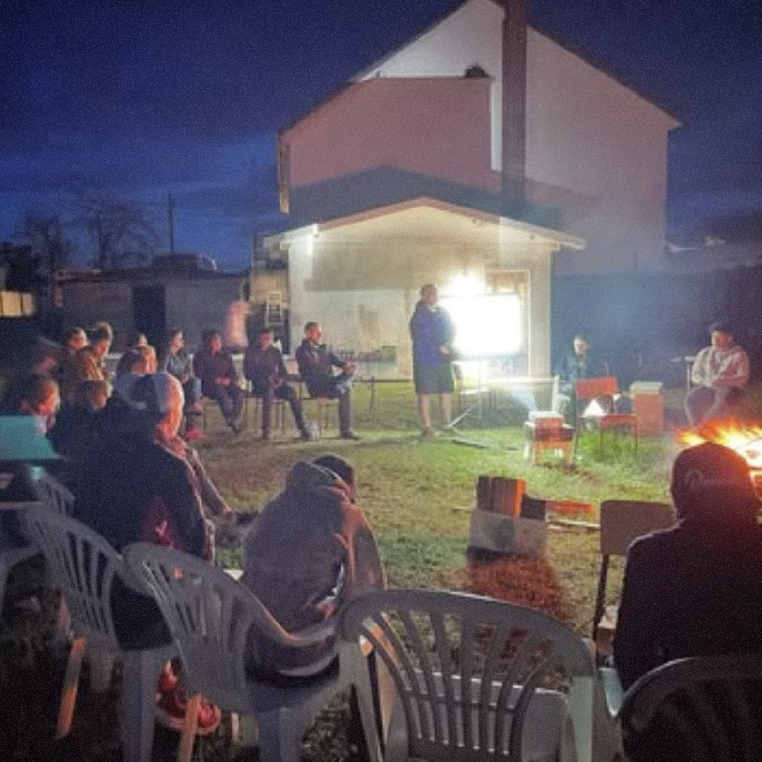 People sitting outdoors in chairs at night, listening to a woman speak in front of a bright light, with a house in the background and a campfire on the right.