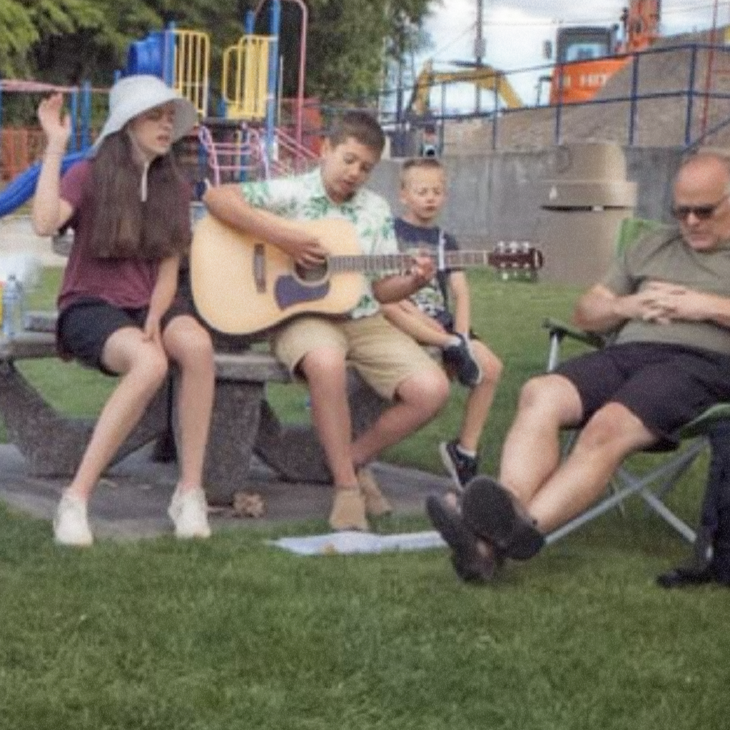 Four people sitting outdoors on a grassy area, with a playground in the background; one person playing guitar, others listening and relaxing.