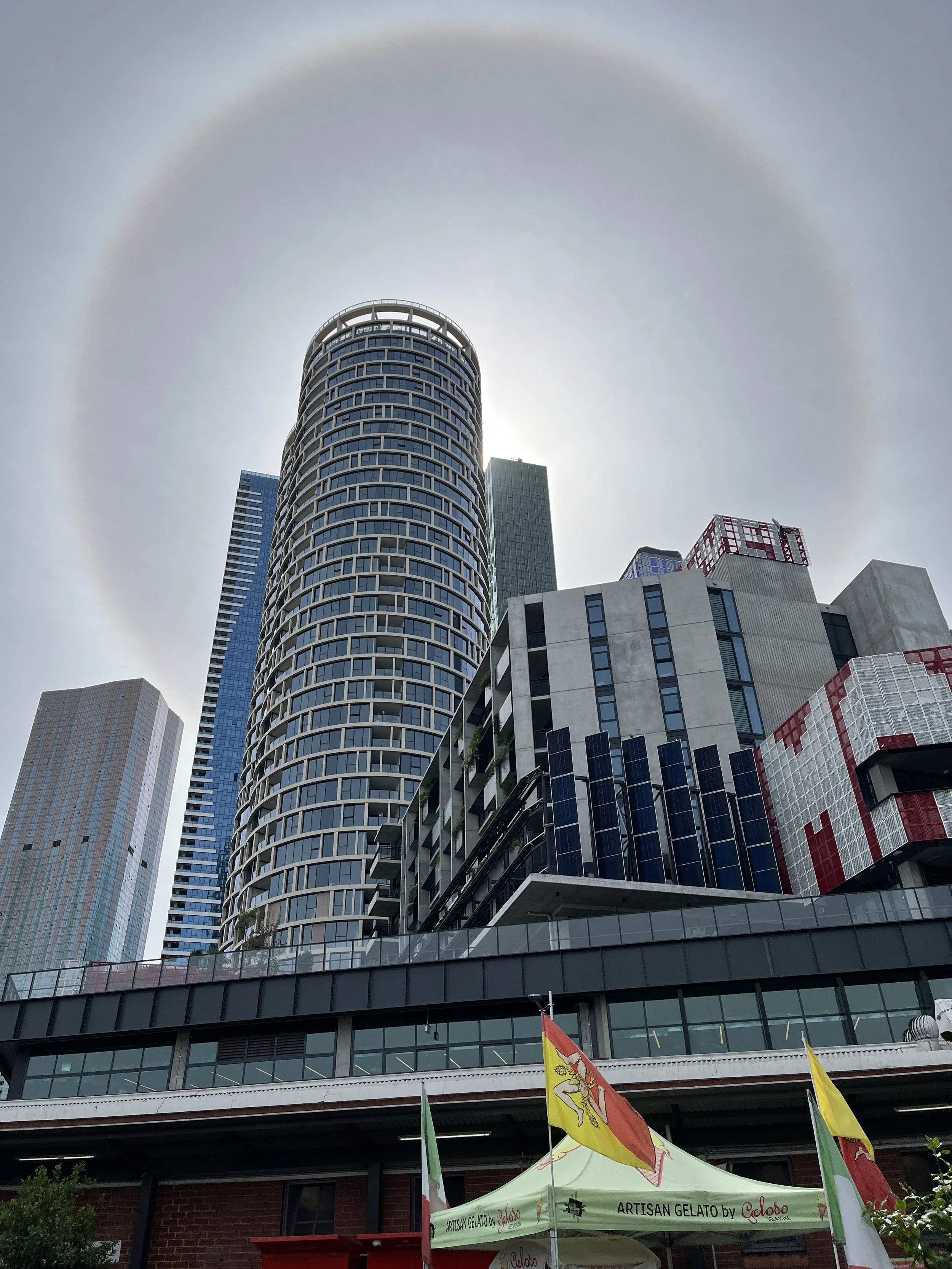 City skyscrapers with a visible halo around the sun behind the tallest building, and a gelato stand with flags in the foreground.