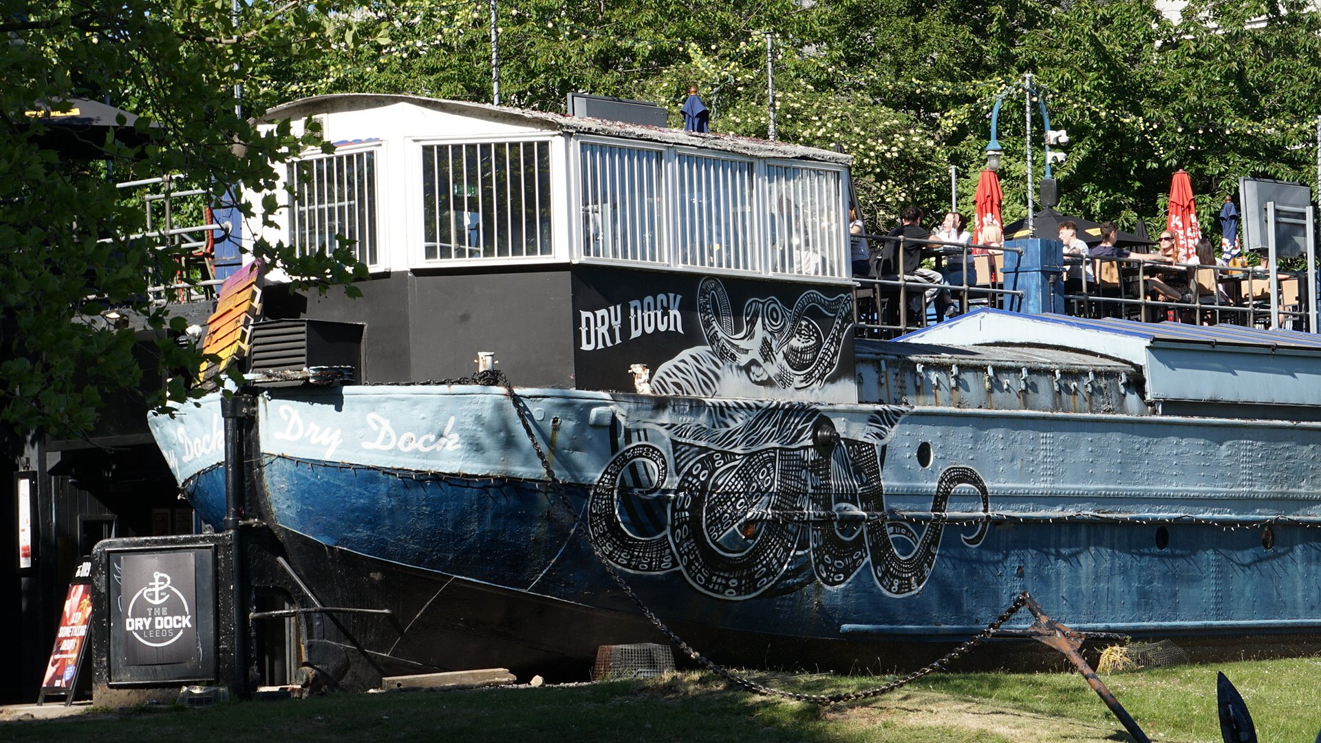 An old boat labeled 'Dry Dock' turned into a bar or restaurant with people dining on an upper deck, surrounded by green trees.