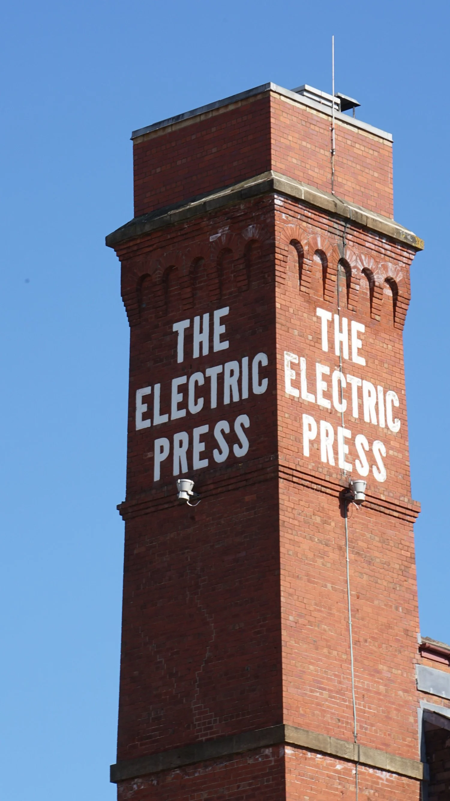 A tall red brick chimney with the words 'THE ELECTRIC PRESS' painted in large white letters on two sides, against a clear blue sky.