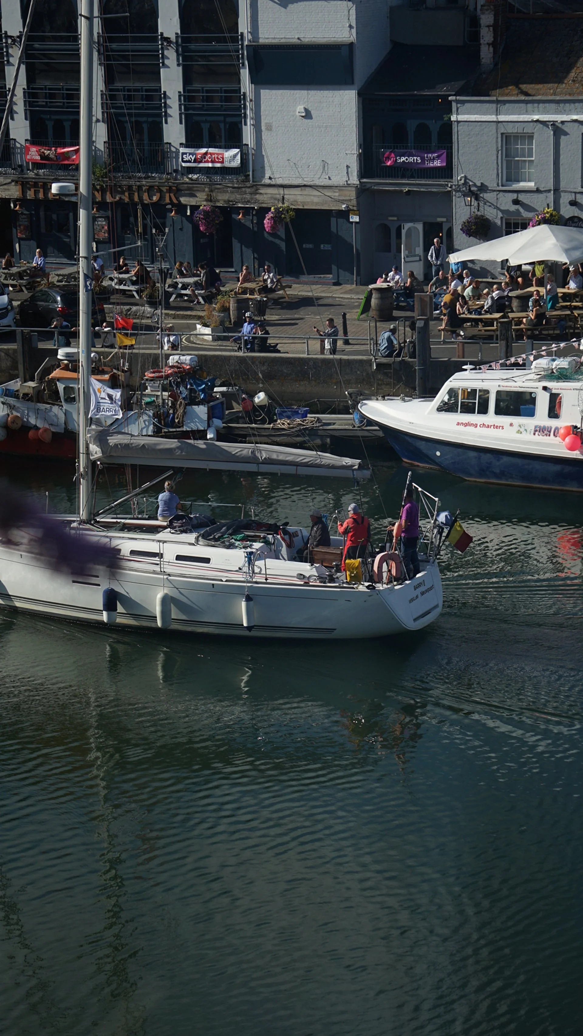 A harbor scene with boats docked, including a sailing boat with people on board and a motorboat named 'FISH ON'. Alongside, there is a promenade with people sitting at tables under umbrellas, some walking, and a building with signs for Sky Sports and